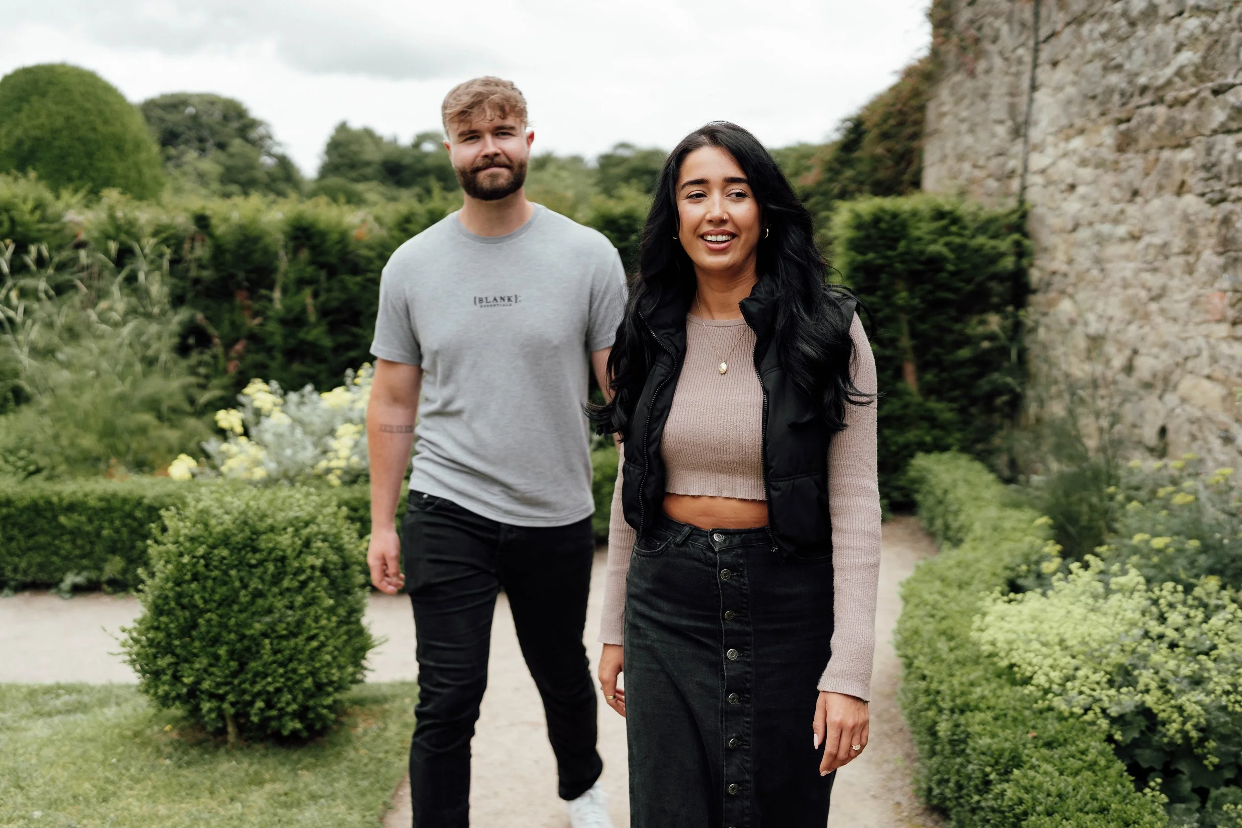 A young woman with long black hair smiling and walking in a garden, a young man with blond hair and beard following behind, surrounded by green bushes and trees, with a stone wall on the right.