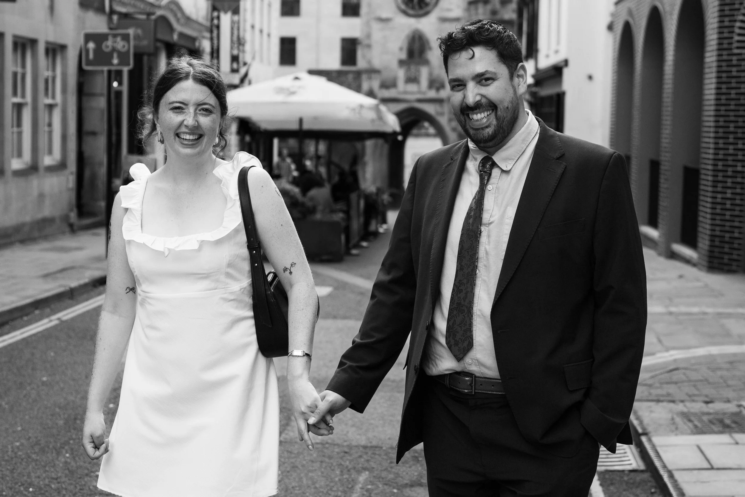 A smiling couple holding hands and walking on a city street, with outdoor dining and buildings in the background, in black and white.