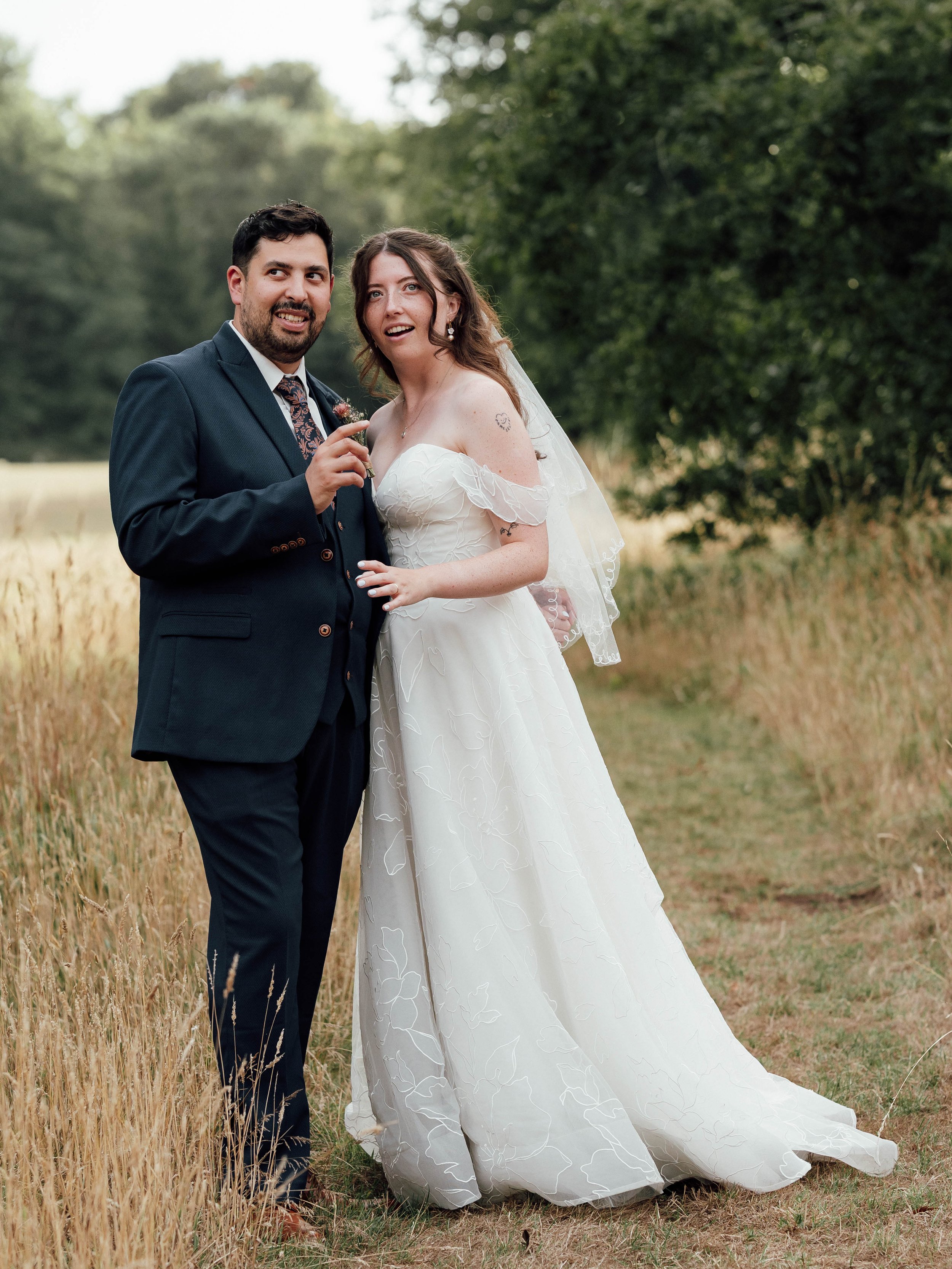 A bride and groom standing outdoors in a field, dressed in wedding attire, with the bride in a white gown and veil and the groom in a dark suit, smiling and posing together.