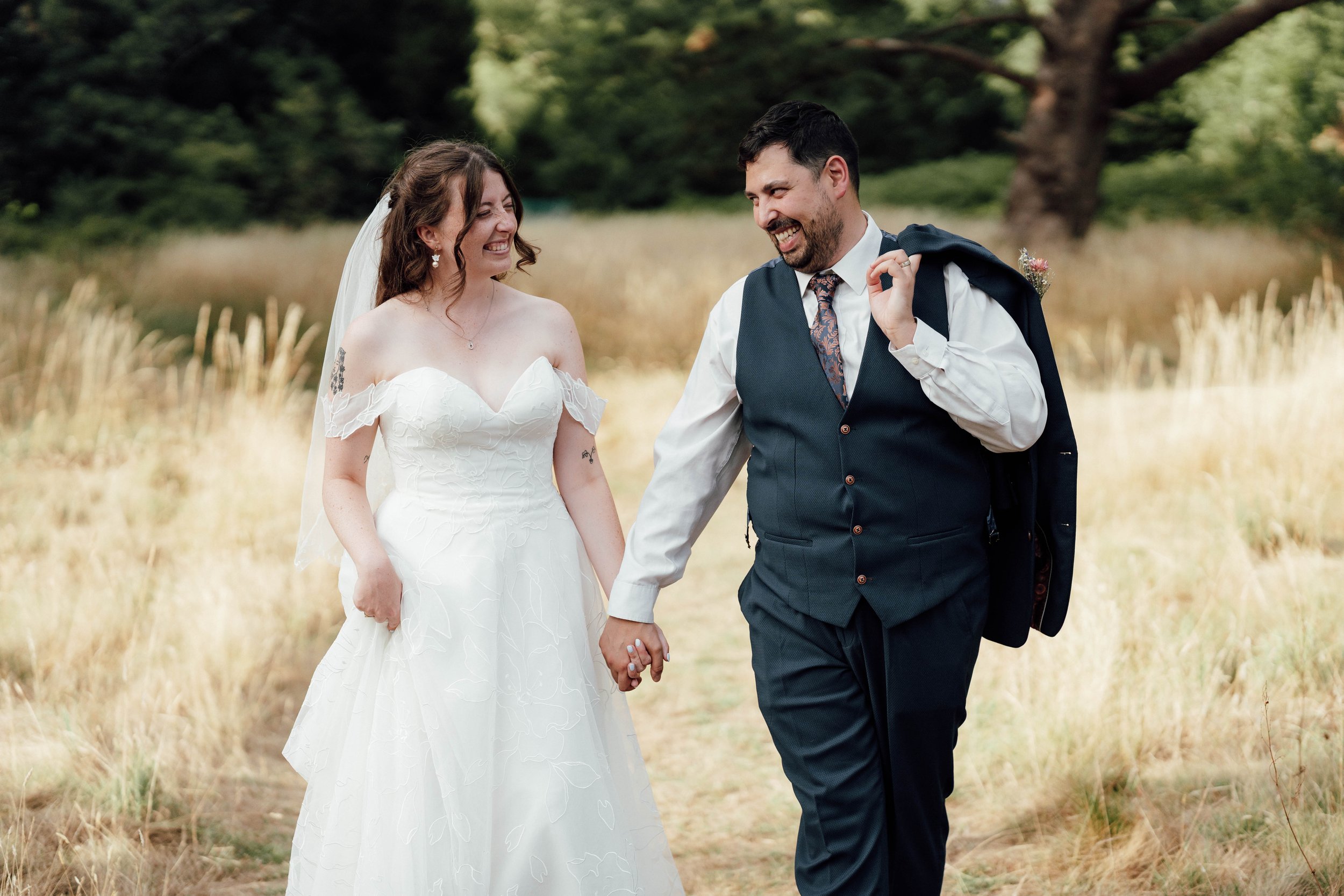 A bride and groom walking hand in hand through a grassy outdoor field, smiling and looking at each other, with trees in the background.