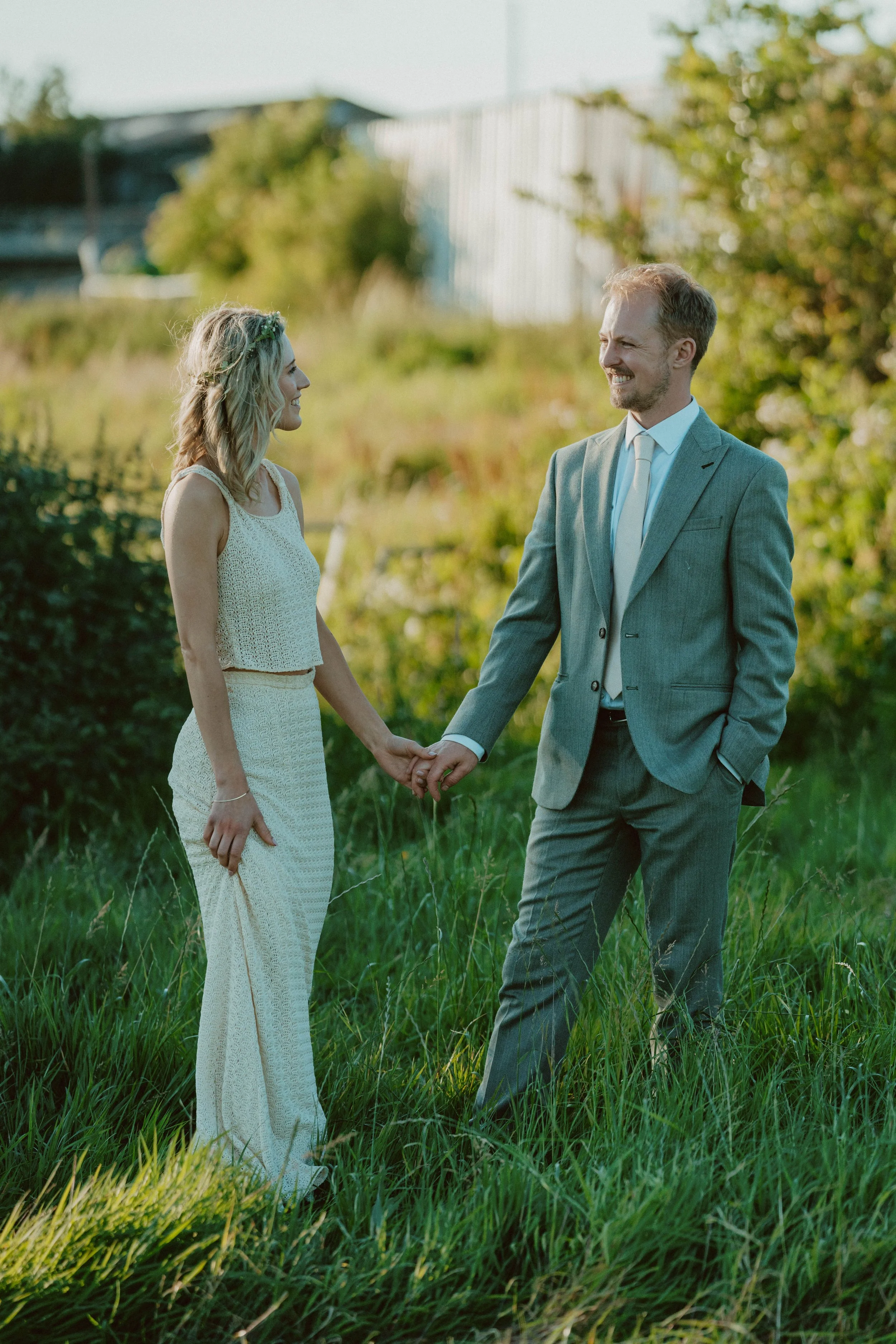 A man in a grey suit and a woman in a white dress holding hands outdoors in a grassy field, smiling at each other.