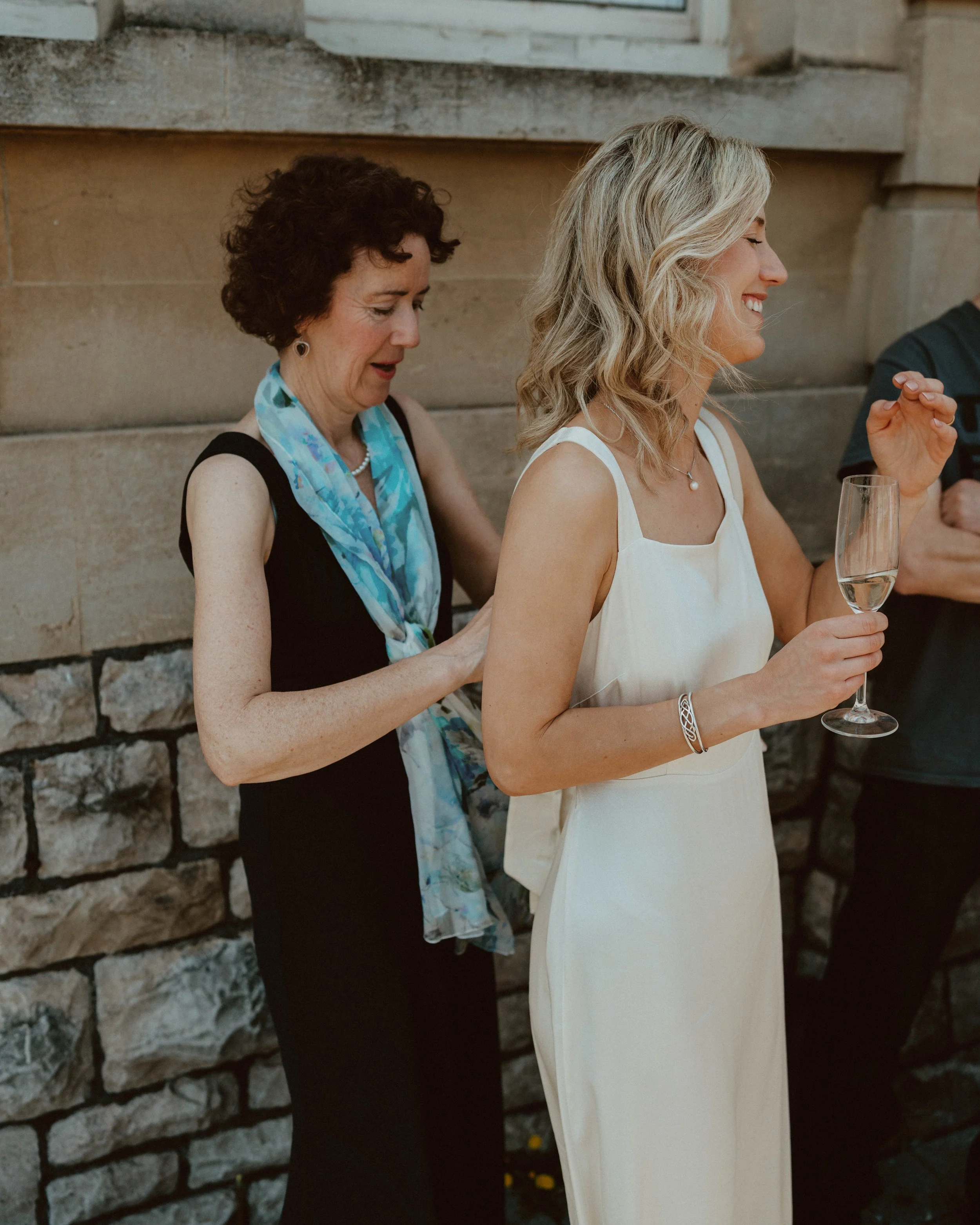 A woman in a white dress holding a glass of champagne, smiling, standing next to a woman in a black dress with a blue scarf, touching her back, against a stone wall.