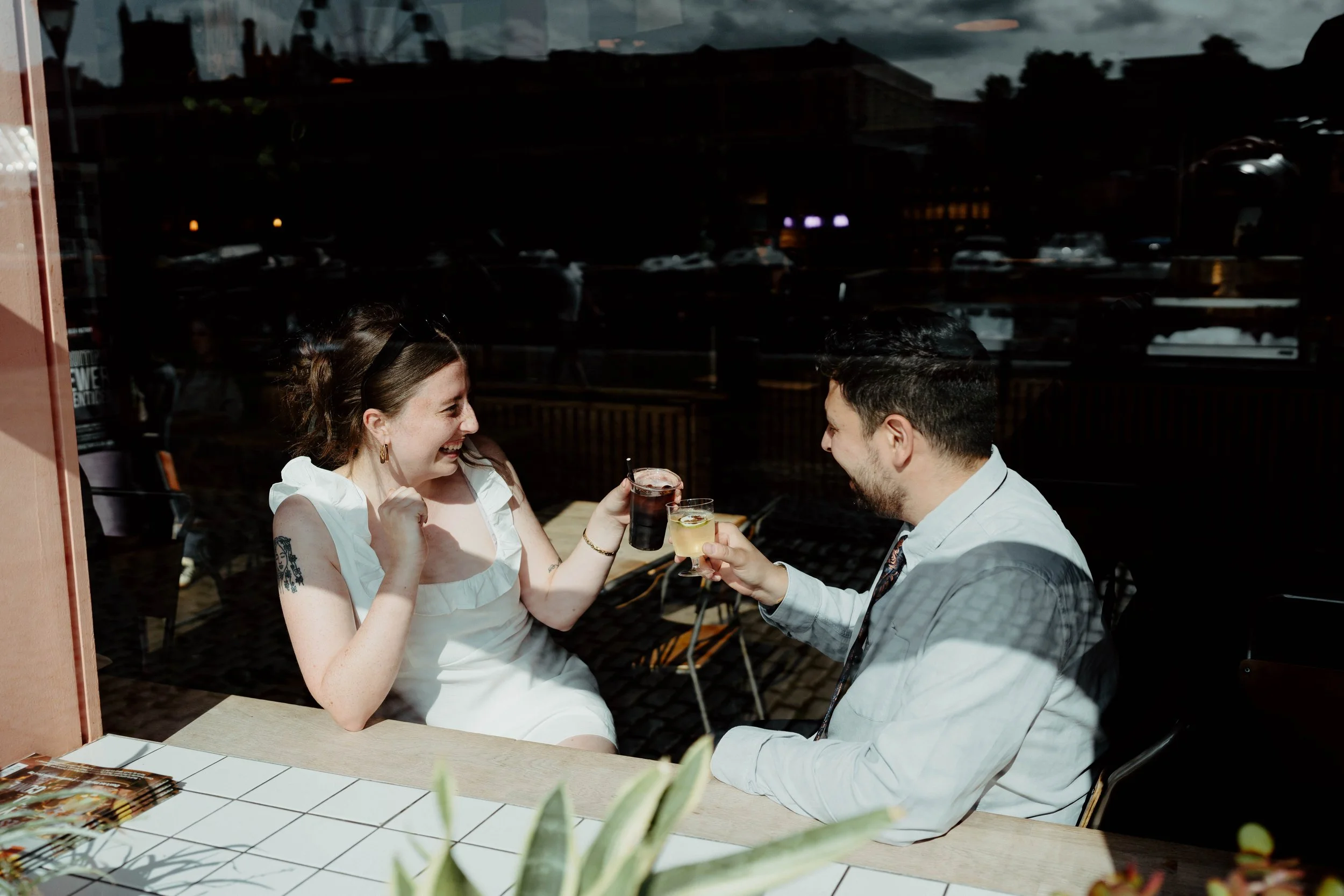 A woman and a man sitting at a table in a restaurant, toasting with drinks and smiling at each other, with a window reflecting the street scene outside.