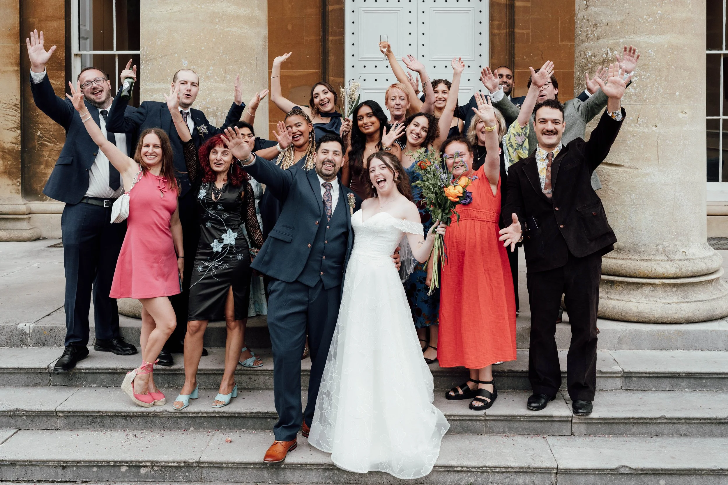 Group of people celebrating a wedding in front of a building with columns, with the bride and groom in the center surrounded by friends and family.