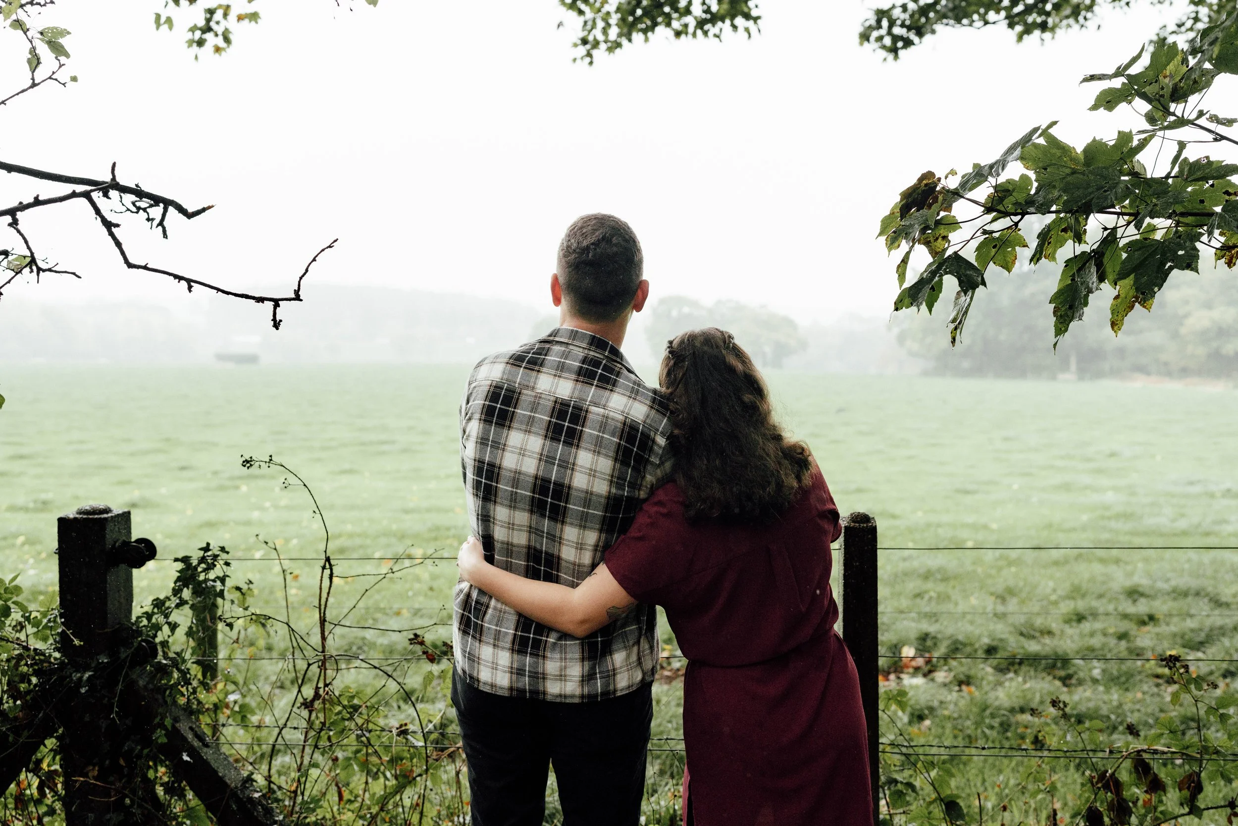A couple standing close together with their backs to the camera, looking out over a green field on a foggy day, with trees and a fence in the foreground.