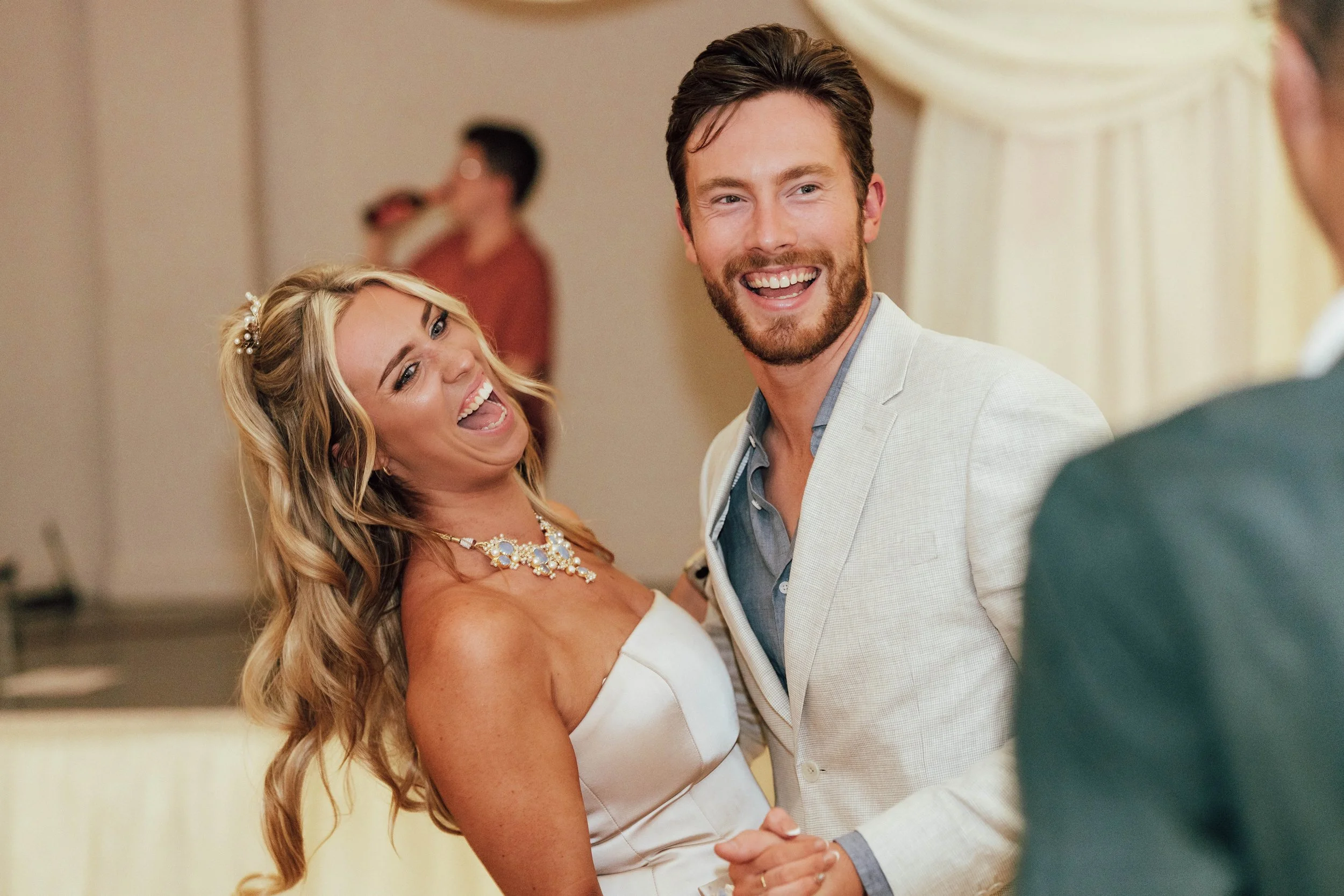 A woman with long blonde hair dancing with a man with brown hair and a beard, both smiling, at a wedding celebration.