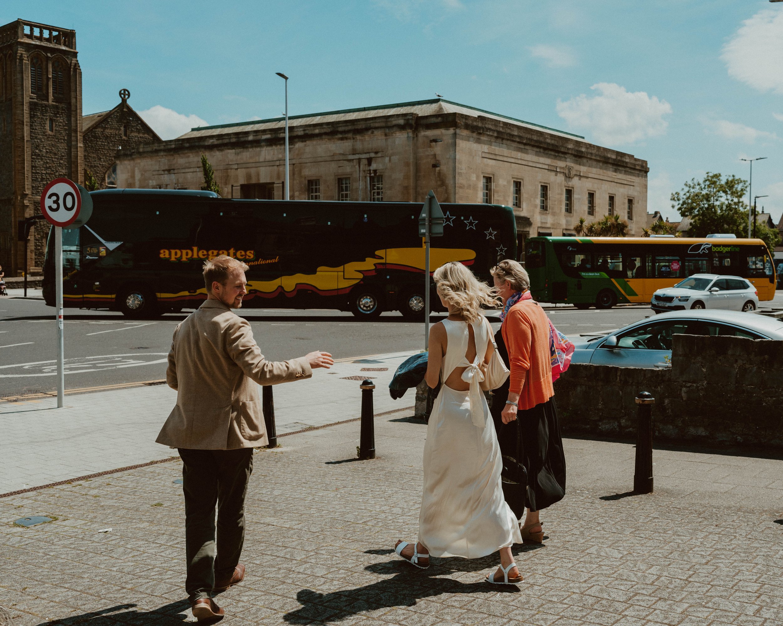 A man gesturing towards two women standing on a city sidewalk with a bus and parked cars in the background during daytime.