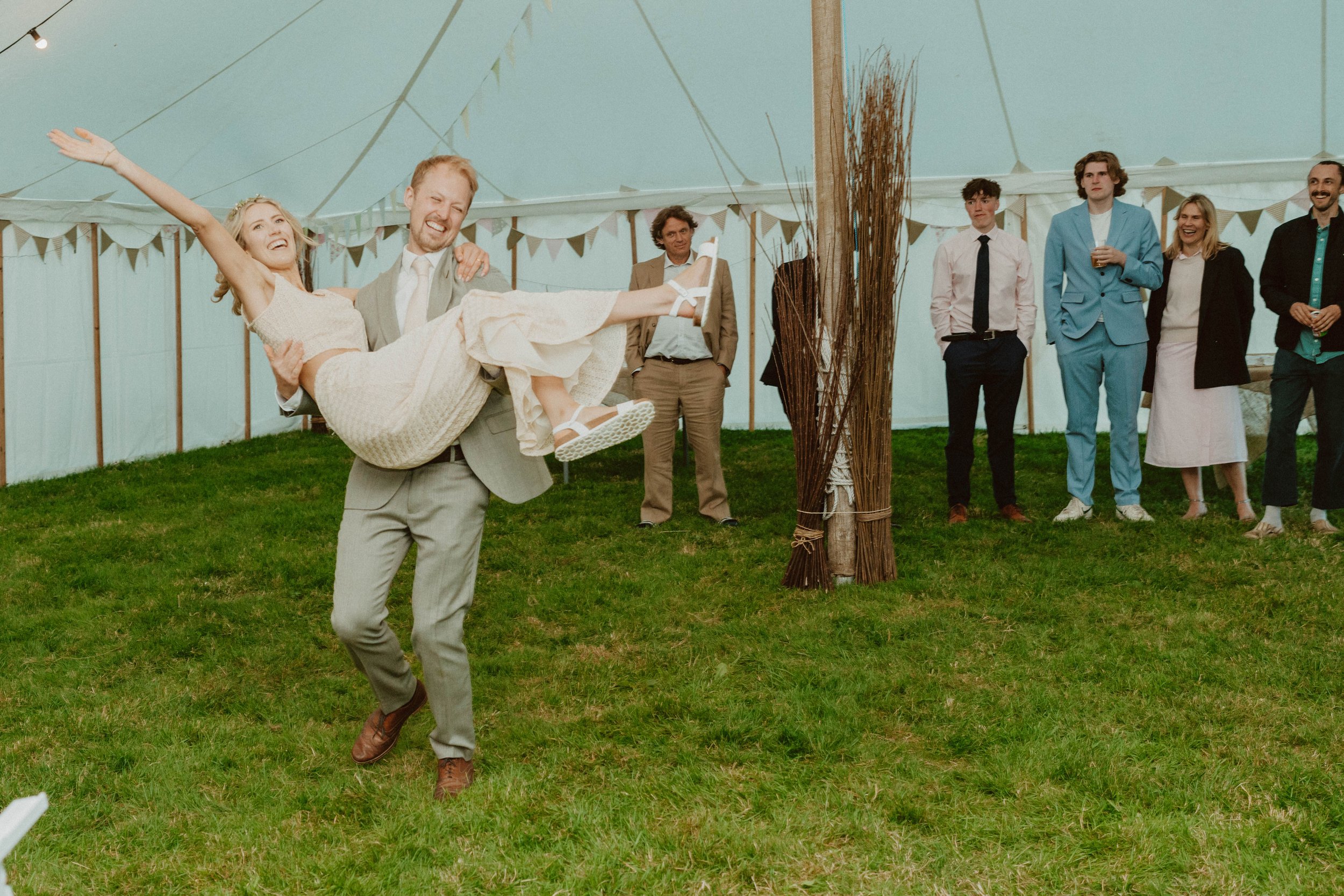 A man in a light gray suit is holding a laughing woman in a cream-colored dress as they dance or celebrate at a gathering under a tent, with other people watching in the background.