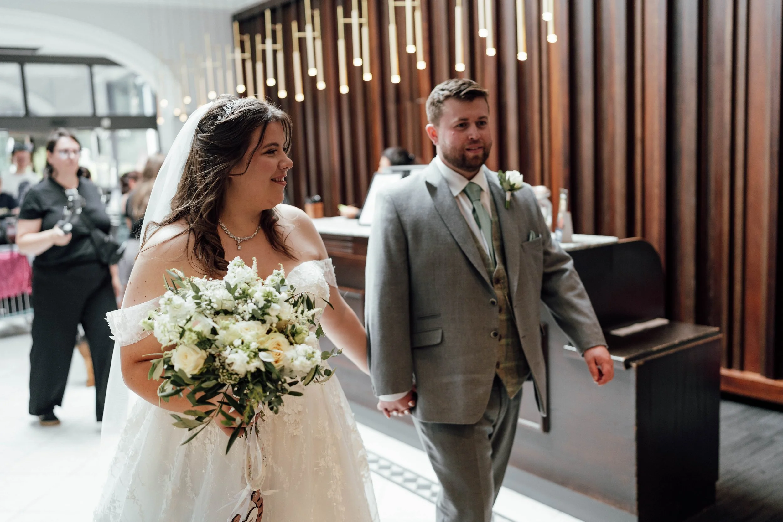 Bride and groom holding hands during wedding ceremony, with bride in white dress holding a bouquet of white flowers, and groom in gray suit with boutonniere, in a venue with wooden wall decor.