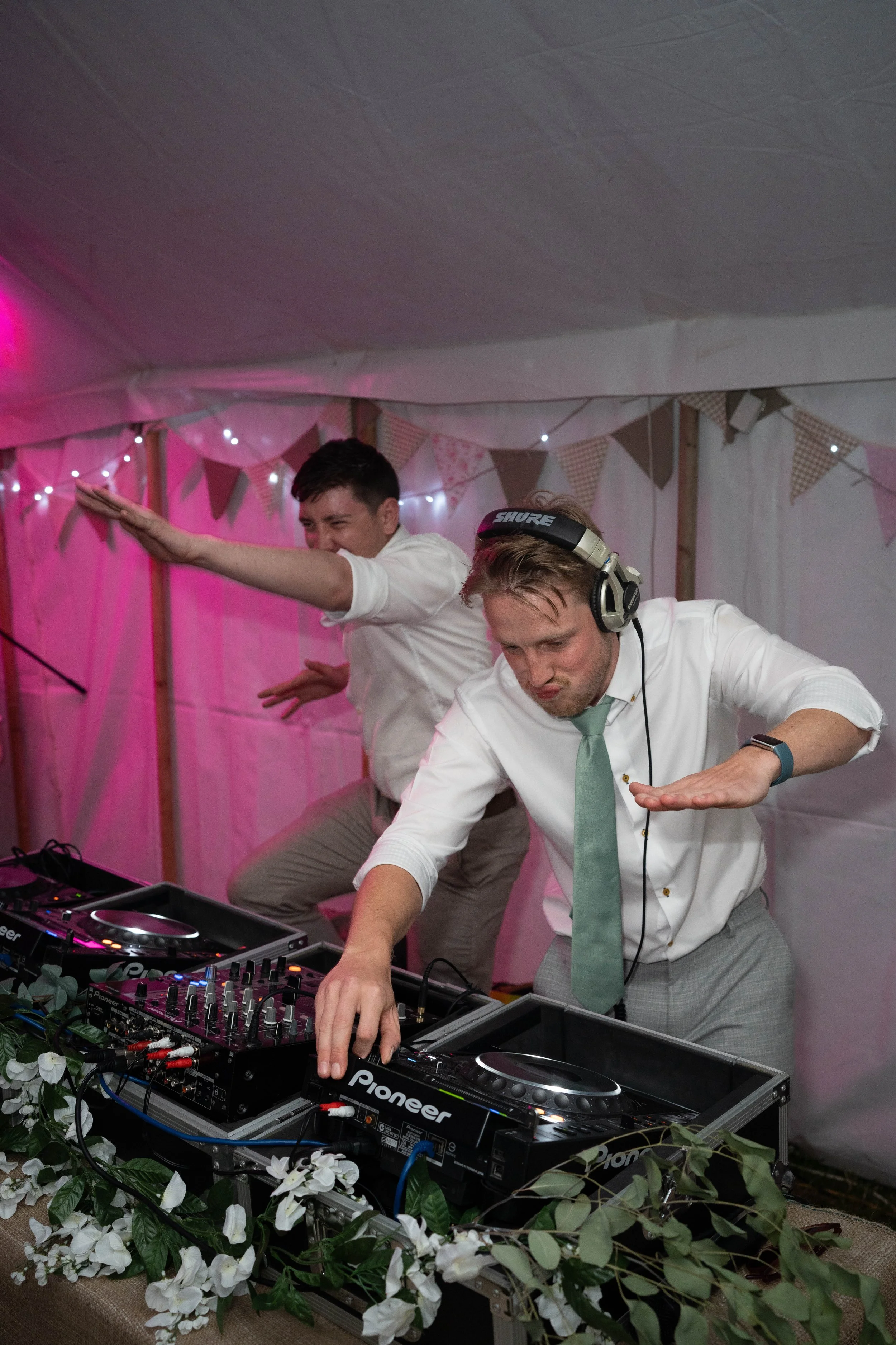 Two men in white shirts and ties, one wearing headphones, dancing and DJing at a decorated event with pink lighting, bunting, and floral decorations.