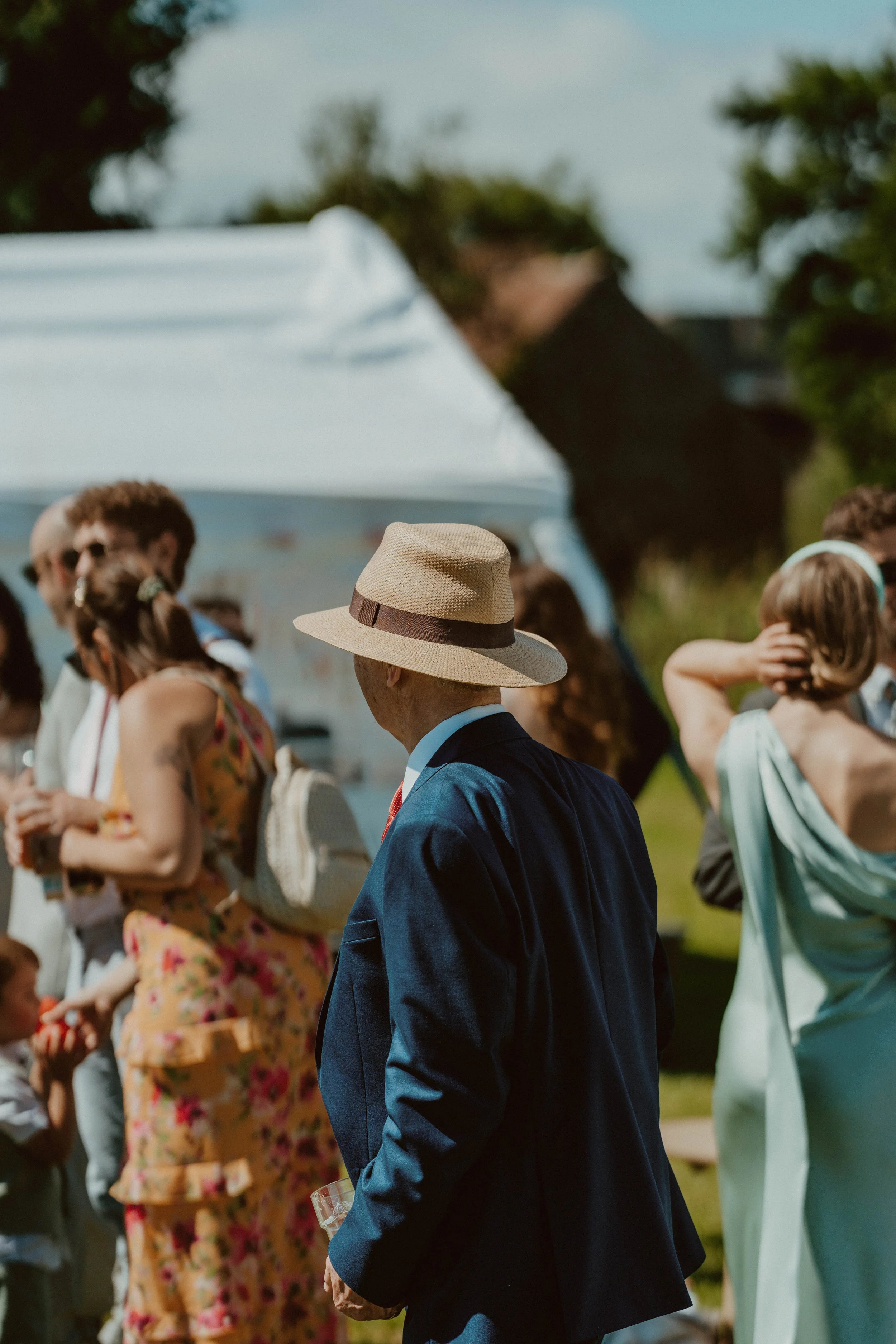 A man in a blue suit and straw hat with a dark band attending an outdoor social gathering with people dressed in summer attire, some wearing hats and sunglasses, and a white tent in the background on a sunny day.