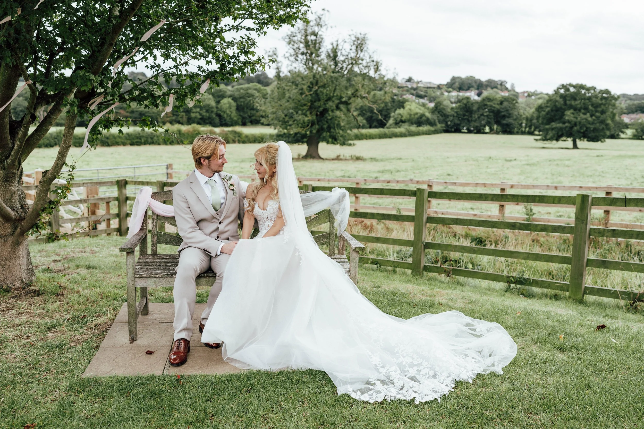 A bride and groom sit on a wooden bench beneath a tree in a green field, gazing at each other. The bride is wearing a white wedding gown with a long train and veil, and the groom is in a light beige suit with a white shirt and tie.