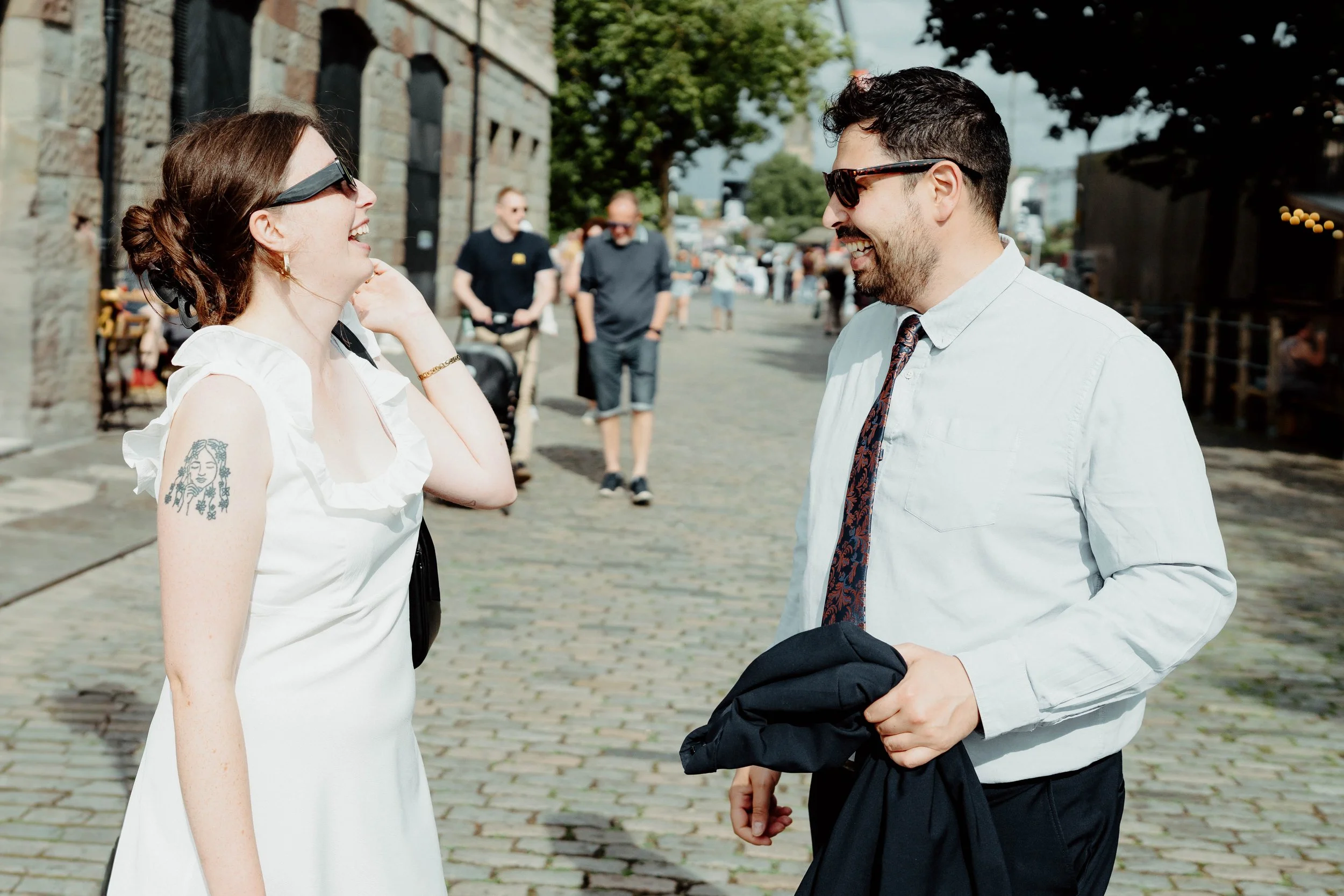 Two people, a woman and a man, are outdoors talking and laughing on a cobblestone street. The woman has brown hair in a bun, wearing sunglasses and a white sleeveless dress, and has a tattoo on her upper left arm. The man has dark hair, beard, sungla