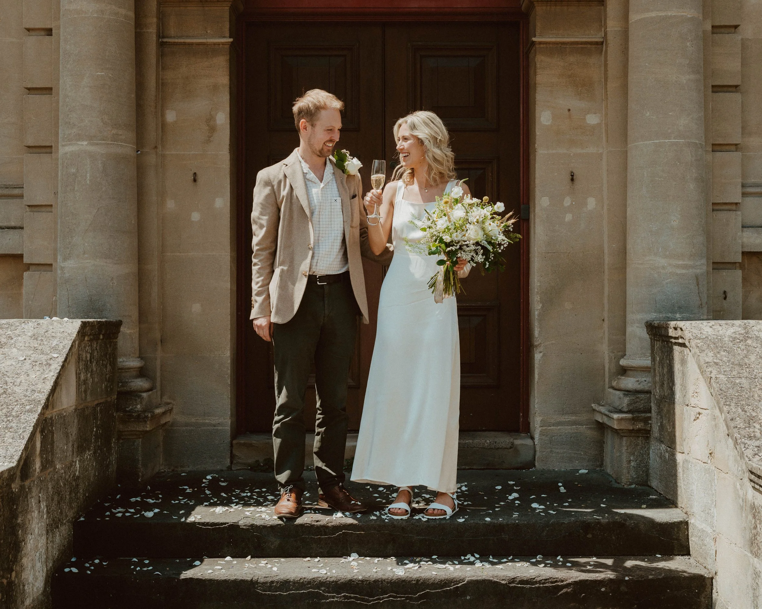 A joyful newlywed couple standing on steps outside a stone building, celebrating with champagne. The bride holds a large bouquet of white flowers and wears a white dress, while the groom wears a beige blazer and dark pants. Petal confetti is scattere