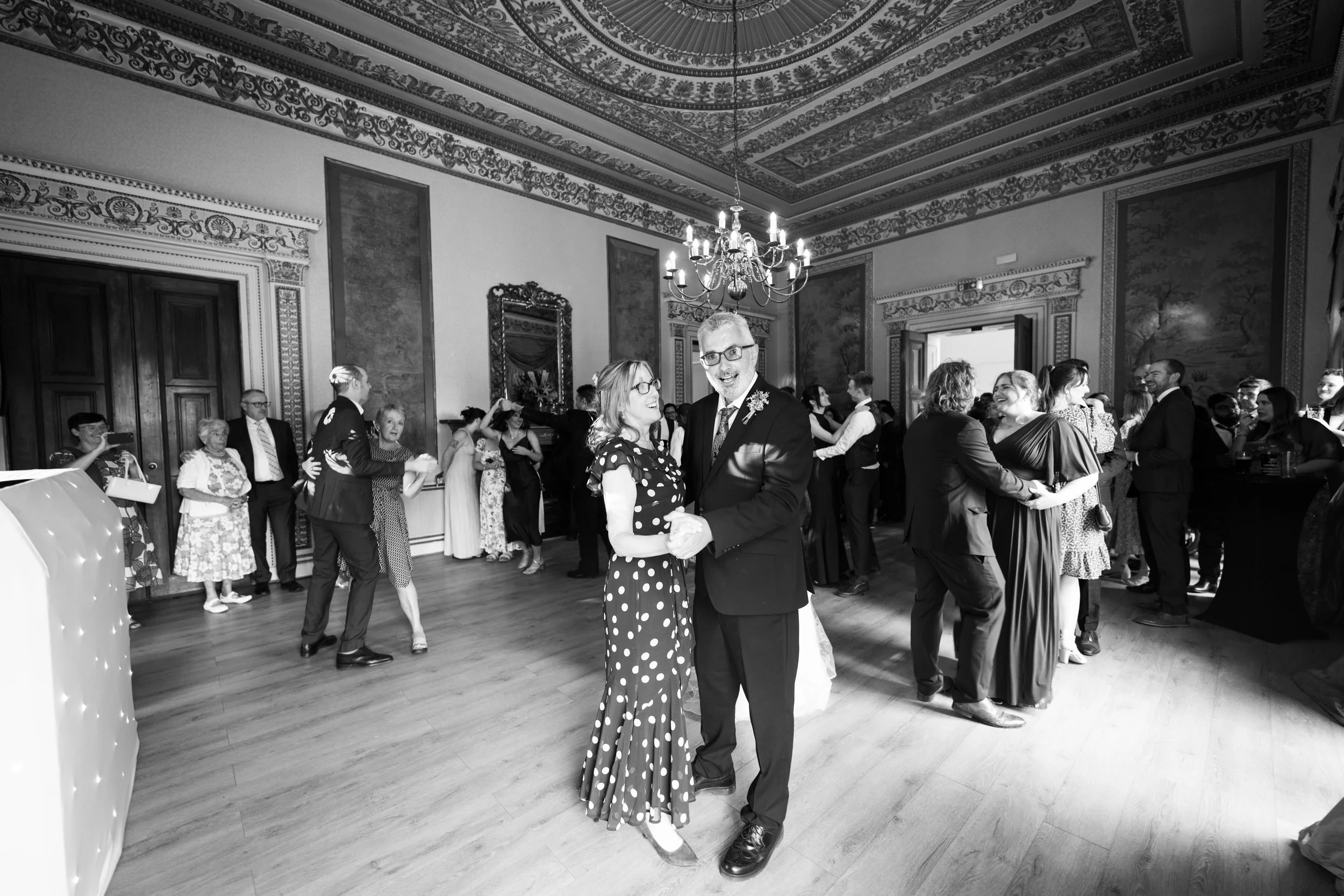 A black-and-white photo of a couple dancing and smiling at a formal event or celebration in a decorated ballroom with other guests dancing and mingling in the background.
