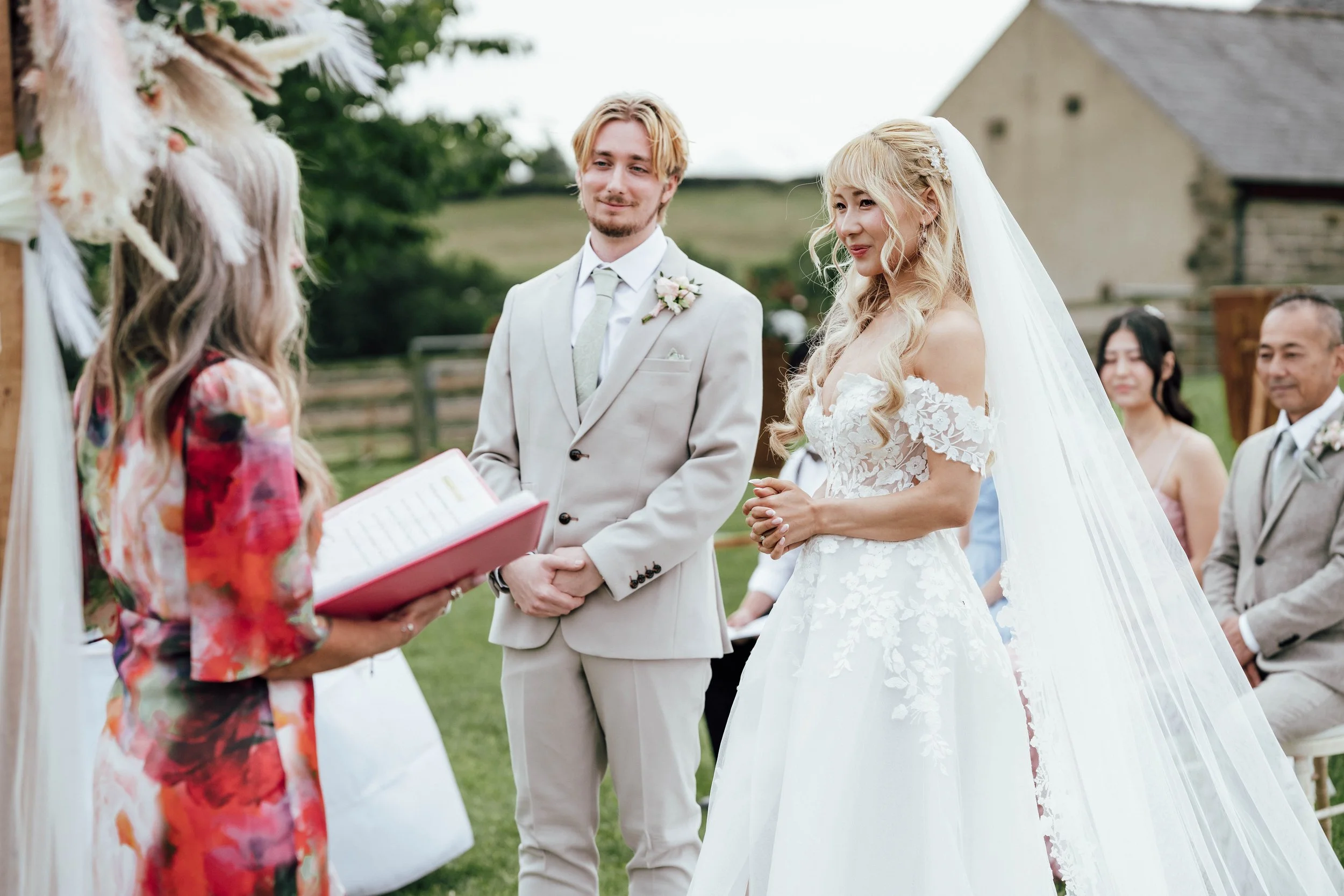 A bride and groom at outdoor wedding ceremony exchanging vows, with a officiant officiating, during daytime, with guests seated in background.