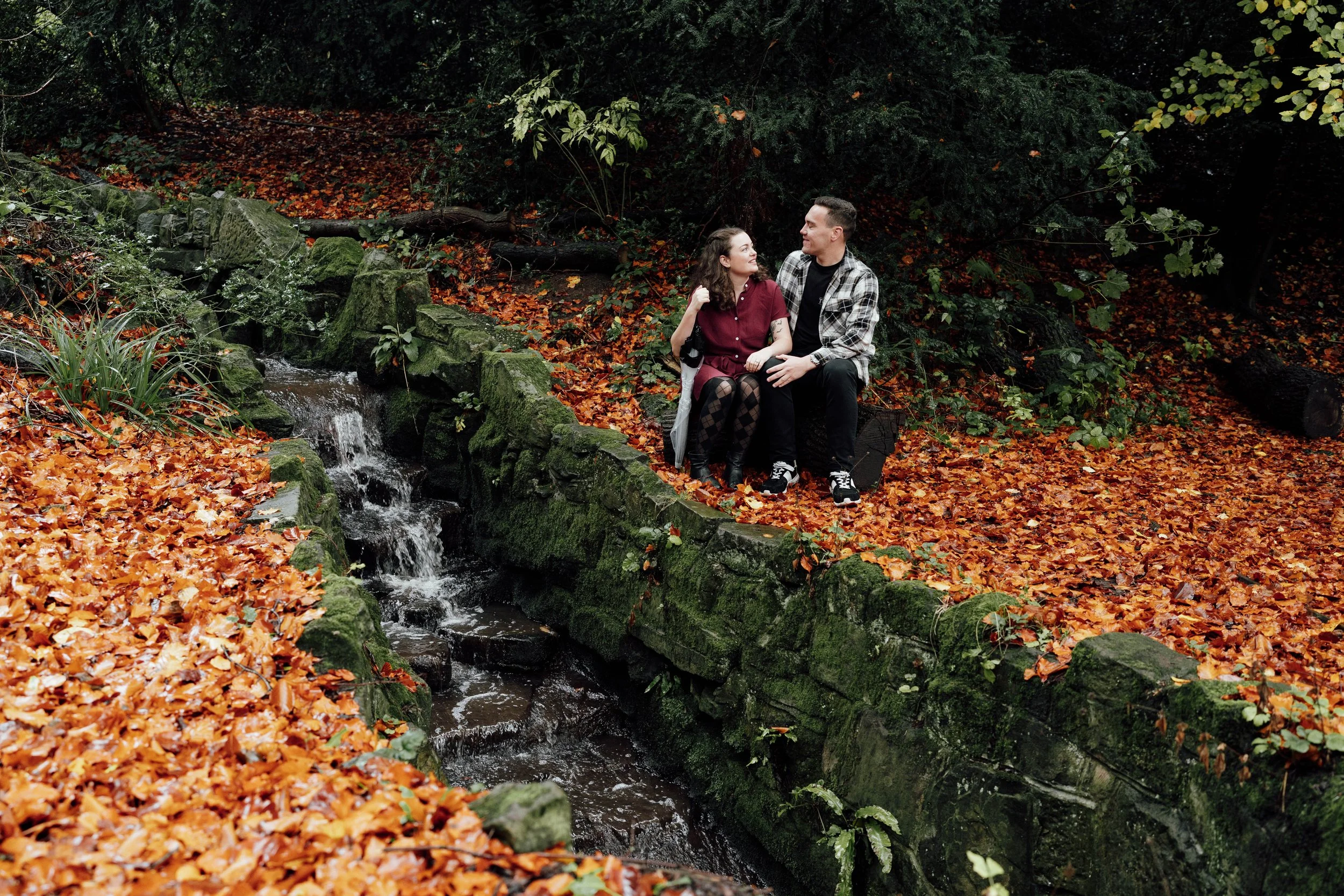 A young couple sitting on a moss-covered stone wall near a small waterfall in a forest with orange leaves on the ground.