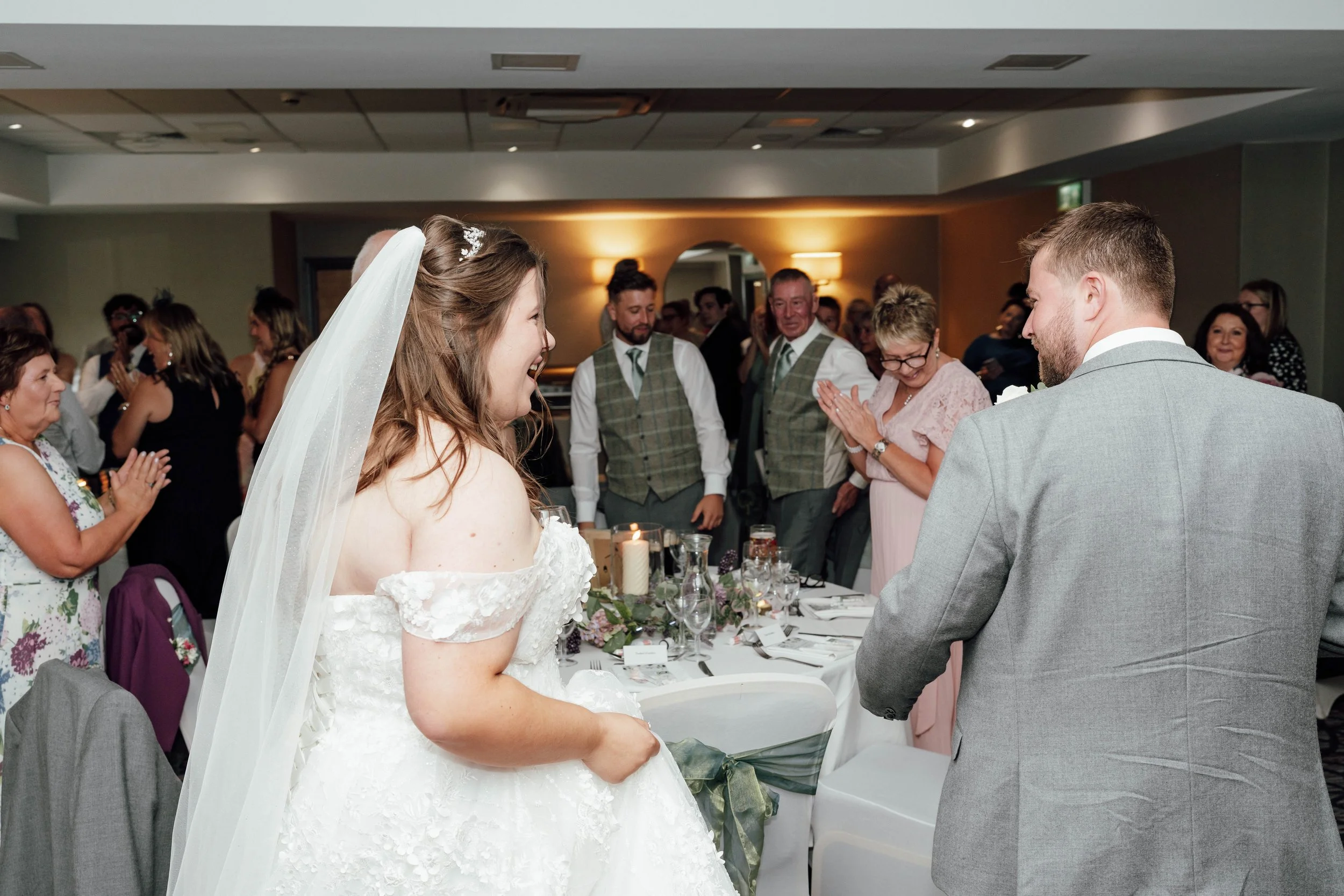 Bride and groom at their wedding reception, surrounded by family and friends, with tables decorated with flowers and candles.
