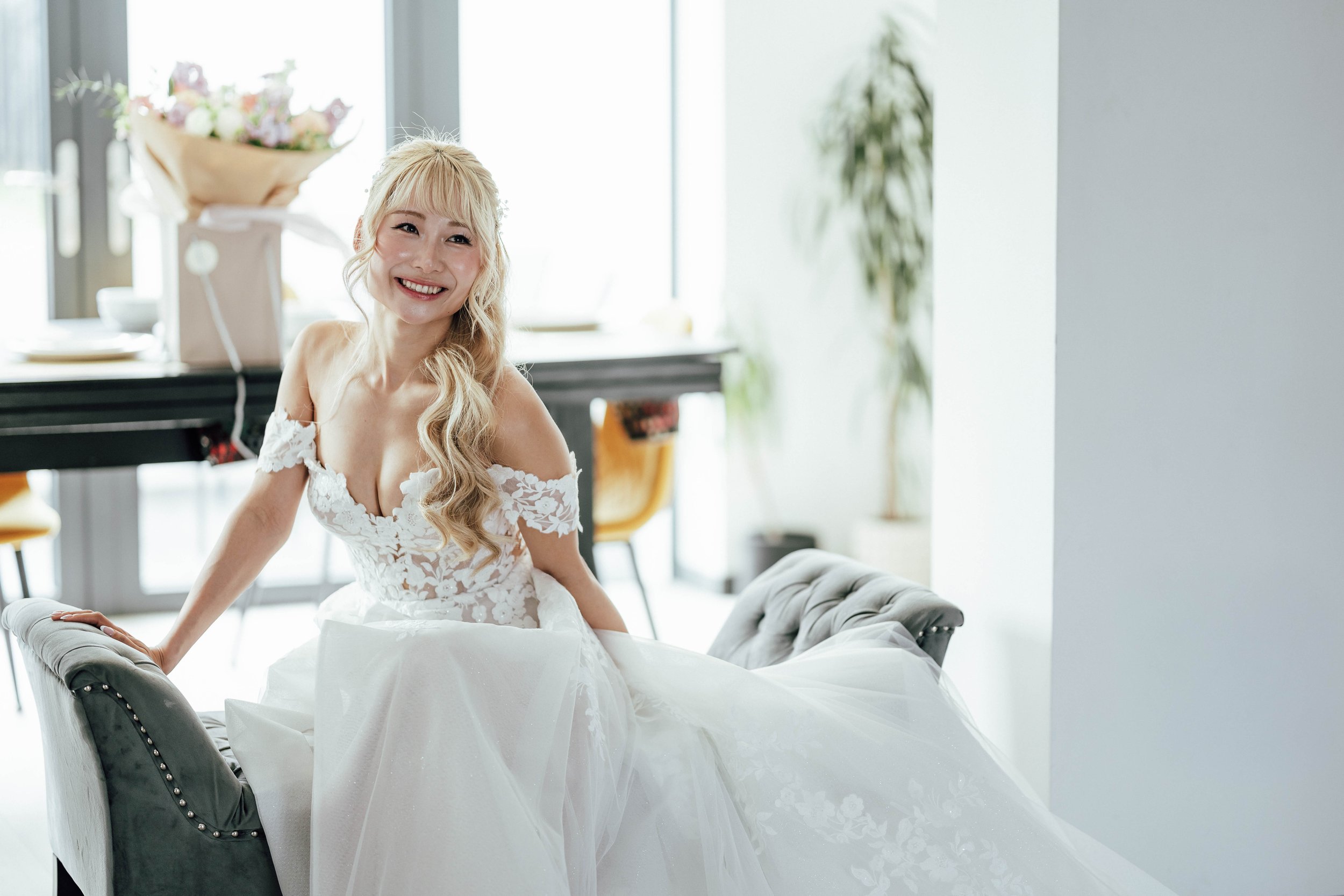 A bride with long blond hair in a white lace wedding dress, sitting on a gray sofa, smiling inside a bright room with large windows and plants.