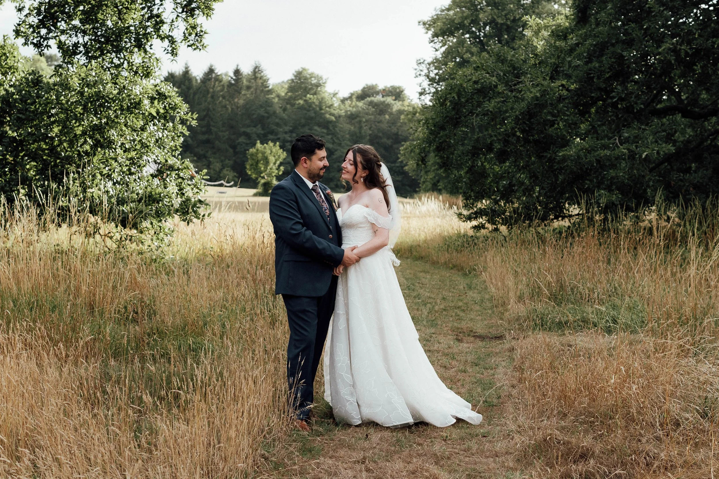 Bride and groom standing in a grassy field holding hands, smiling at each other during wedding photos, surrounded by trees and nature.
