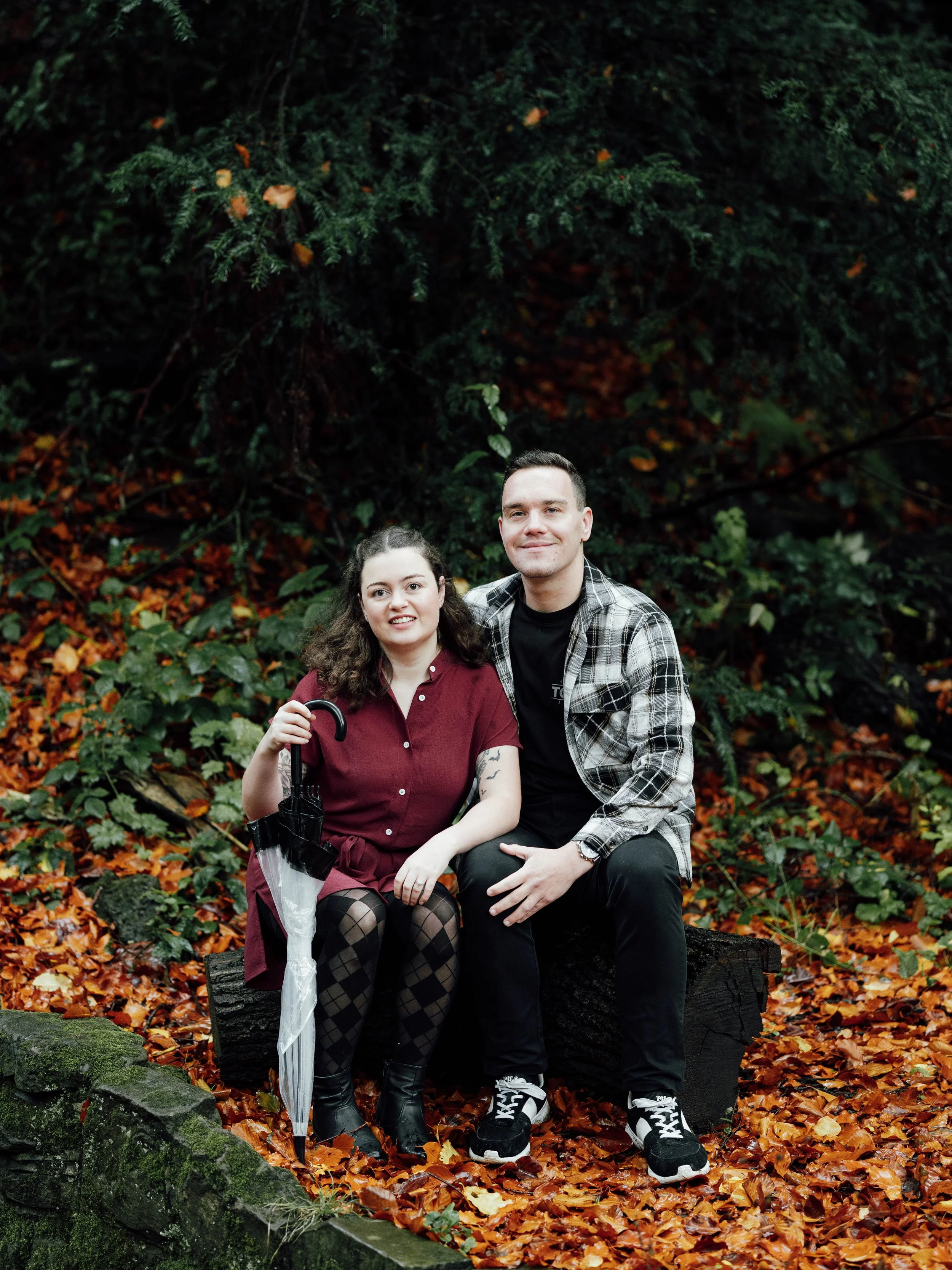 A young woman sitting on a log in a forest with fallen leaves, holding an umbrella, and a young man sitting beside her. They are both smiling and posing for the photo.