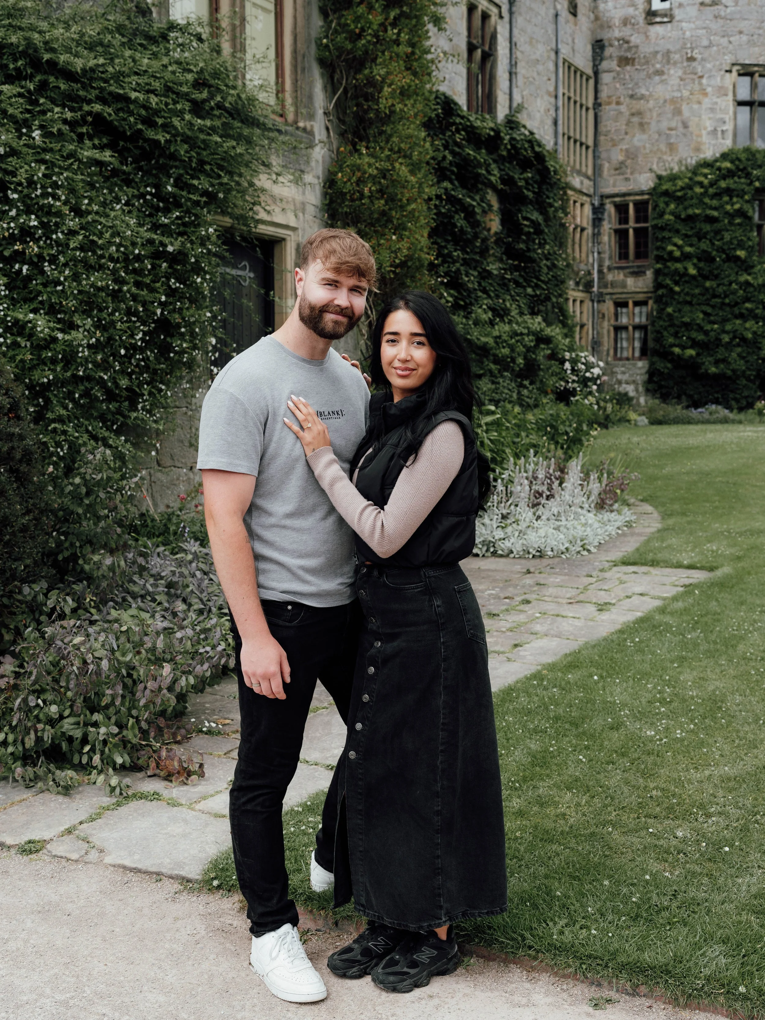 A happy couple standing outdoors on a stone pathway in front of an old stone building with windows and green foliage.