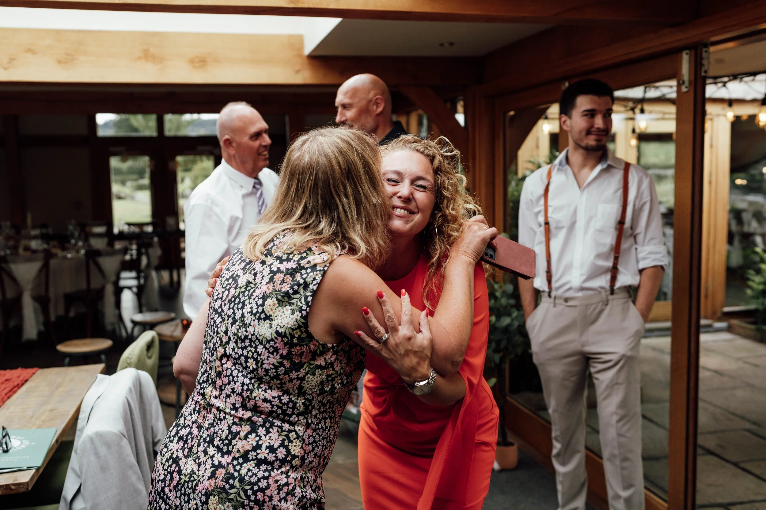 Two women embrace at a celebration, one smiling with eyes closed, holding a phone, with three men standing in the background inside a wooden structure.