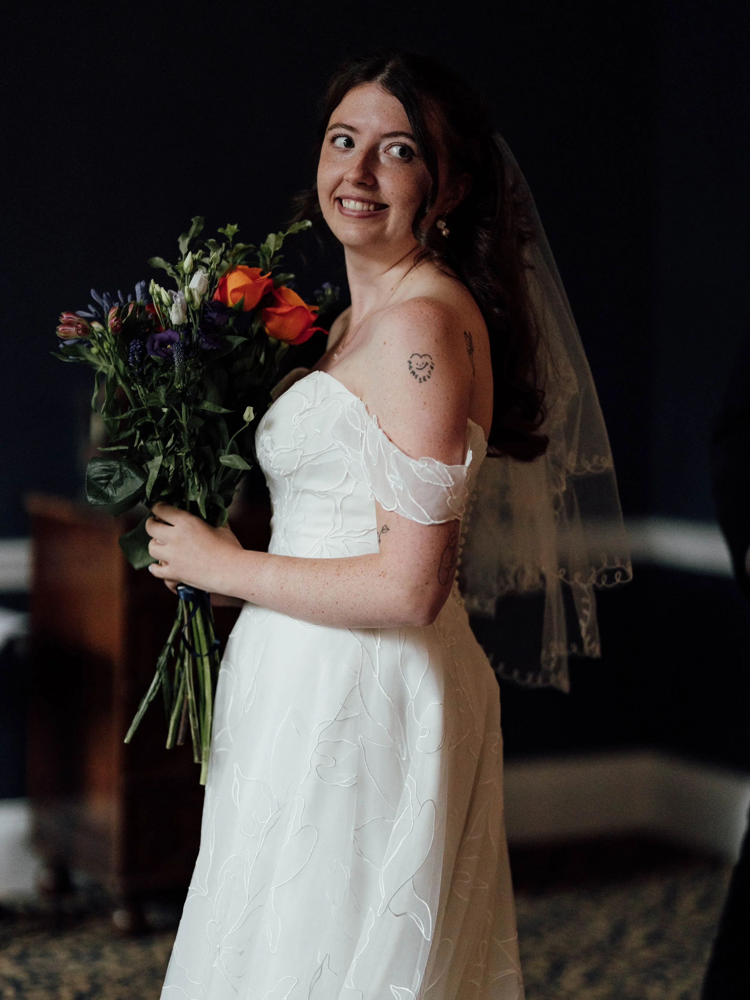 A smiling bride in a white wedding dress holding a bouquet of flowers indoors with a dark background.