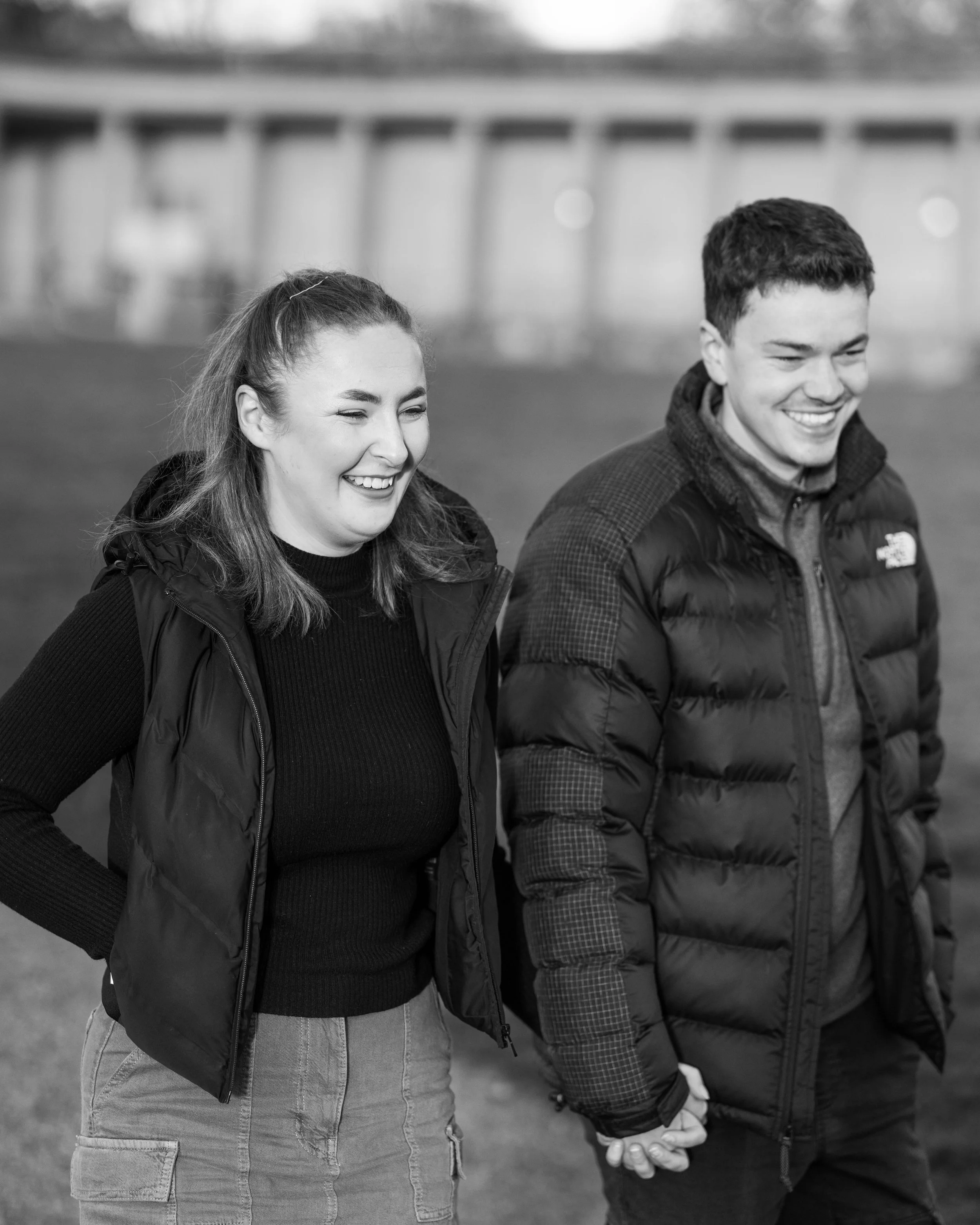 A smiling young woman and man holding hands outdoors, dressed in warm jackets, with a blurred bridge in the background.