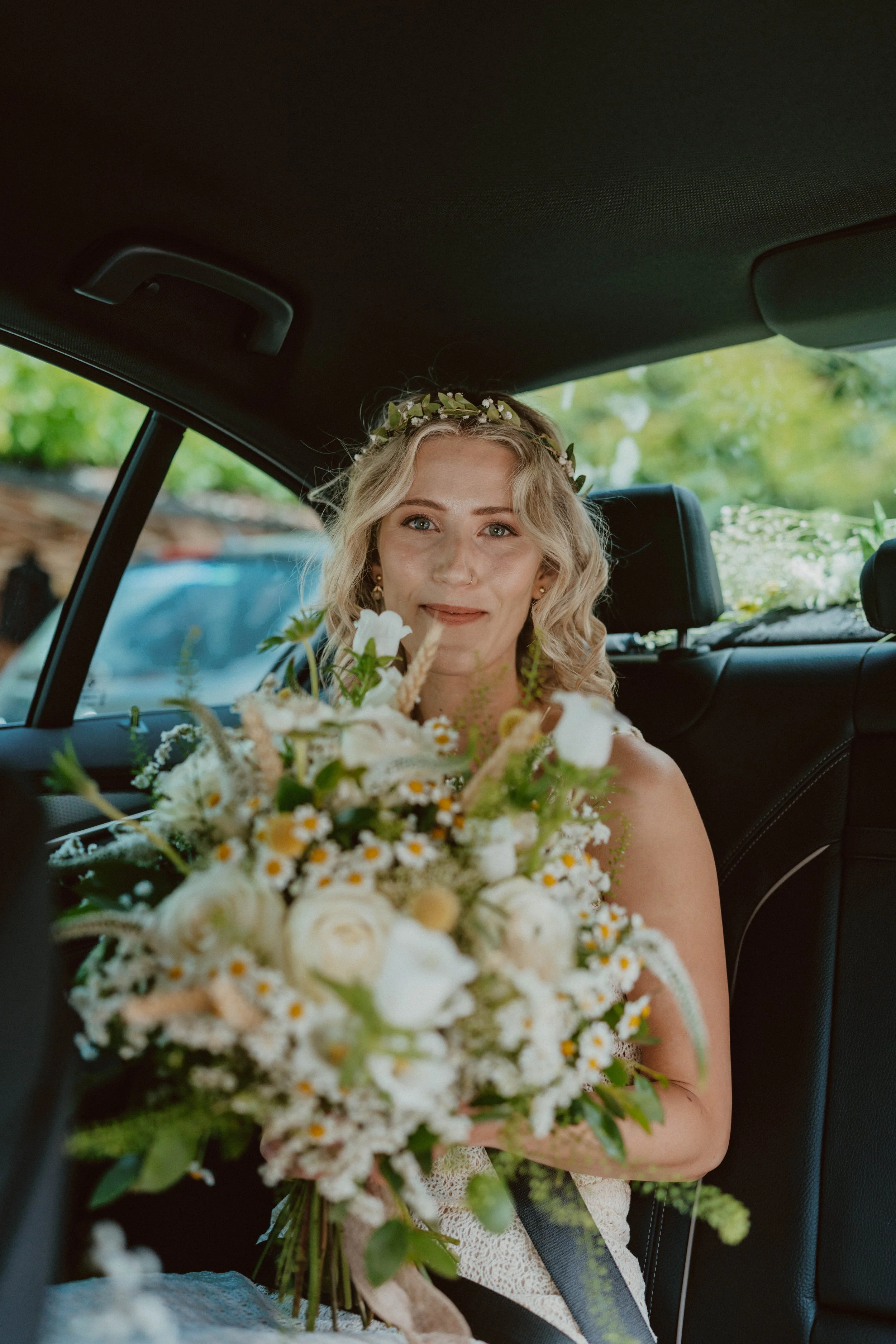 Bride sitting in the backseat of a car holding a large bouquet of white flowers, wearing a floral crown, with a happy expression.