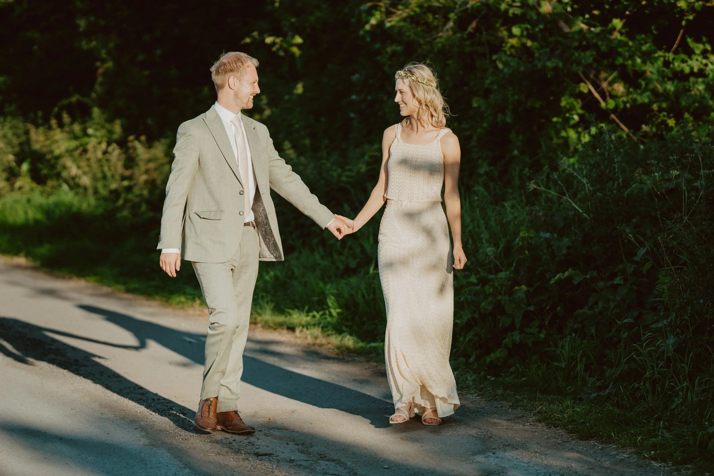 A man and woman holding hands and walking along a dirt path in a wooded area during daytime. The man is wearing a light-colored suit, and the woman is in a long, sleeveless dress.