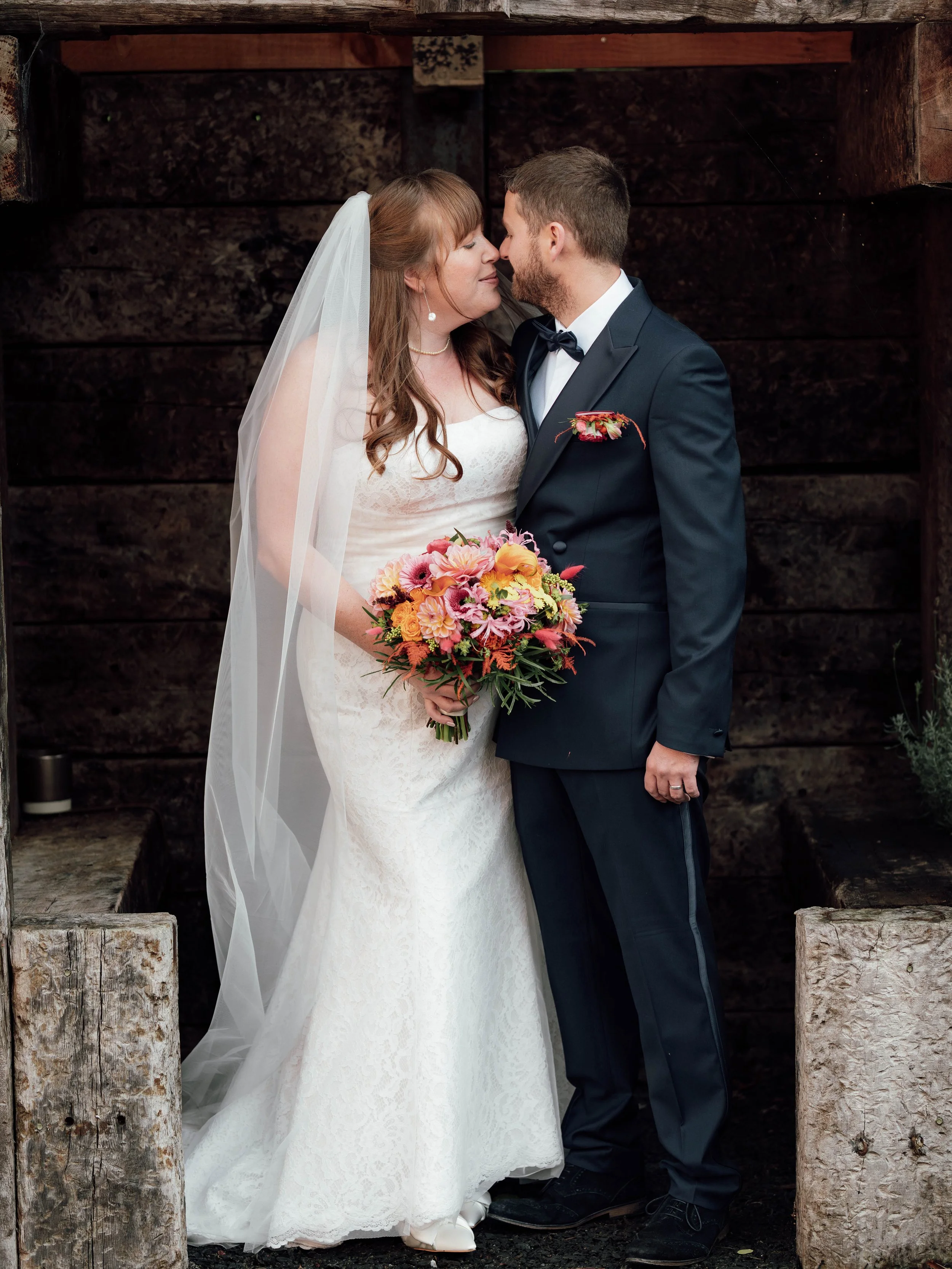 Bride and groom in wedding attire face each other, with the bride holding a colorful bouquet, standing in front of a dark wooden background.