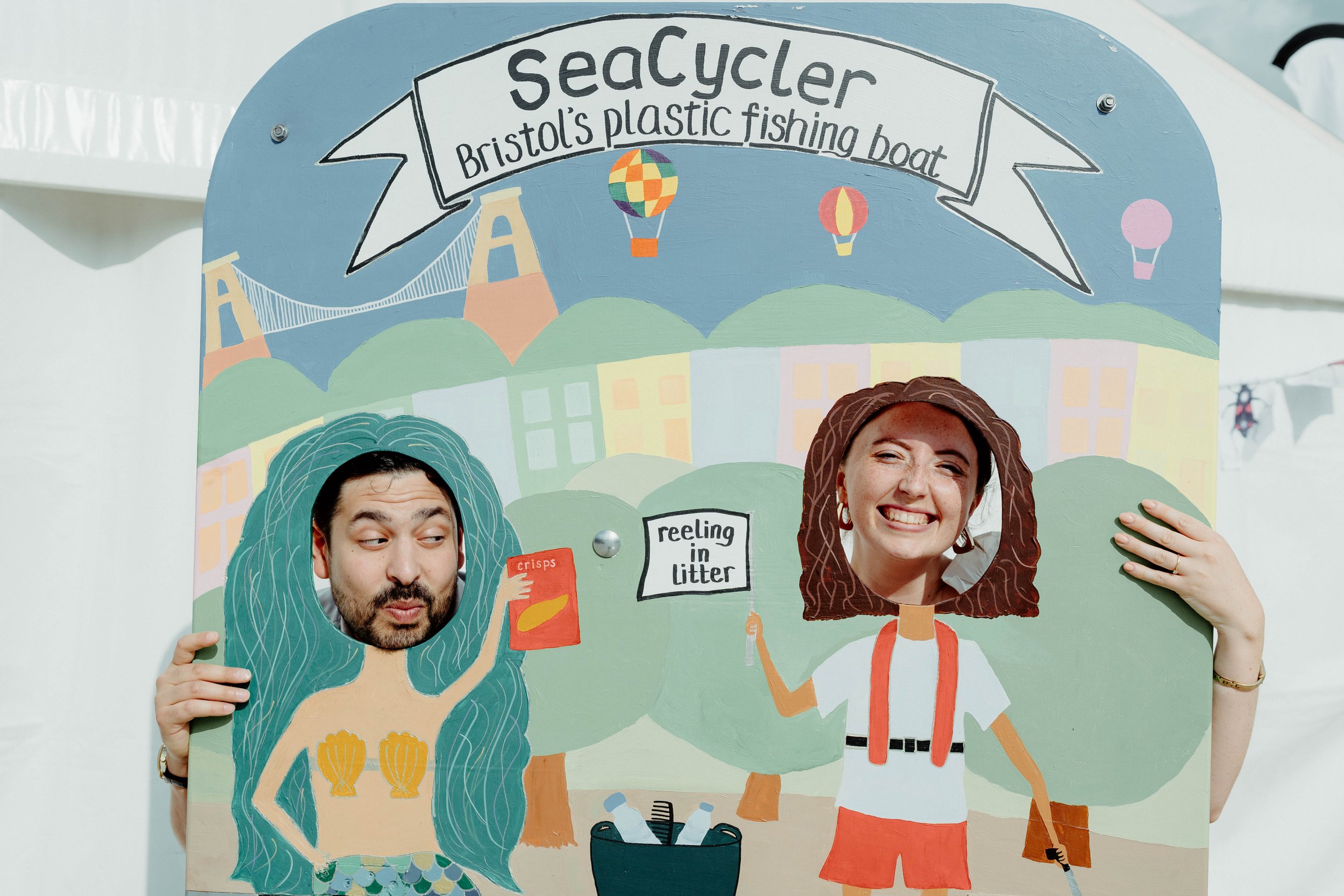Two people with cutout faces posing through a painted cardboard scenery at a Bristol Seacycler event, featuring a mermaid with a snack and a person holding a sign that says 'reeling in litter'.