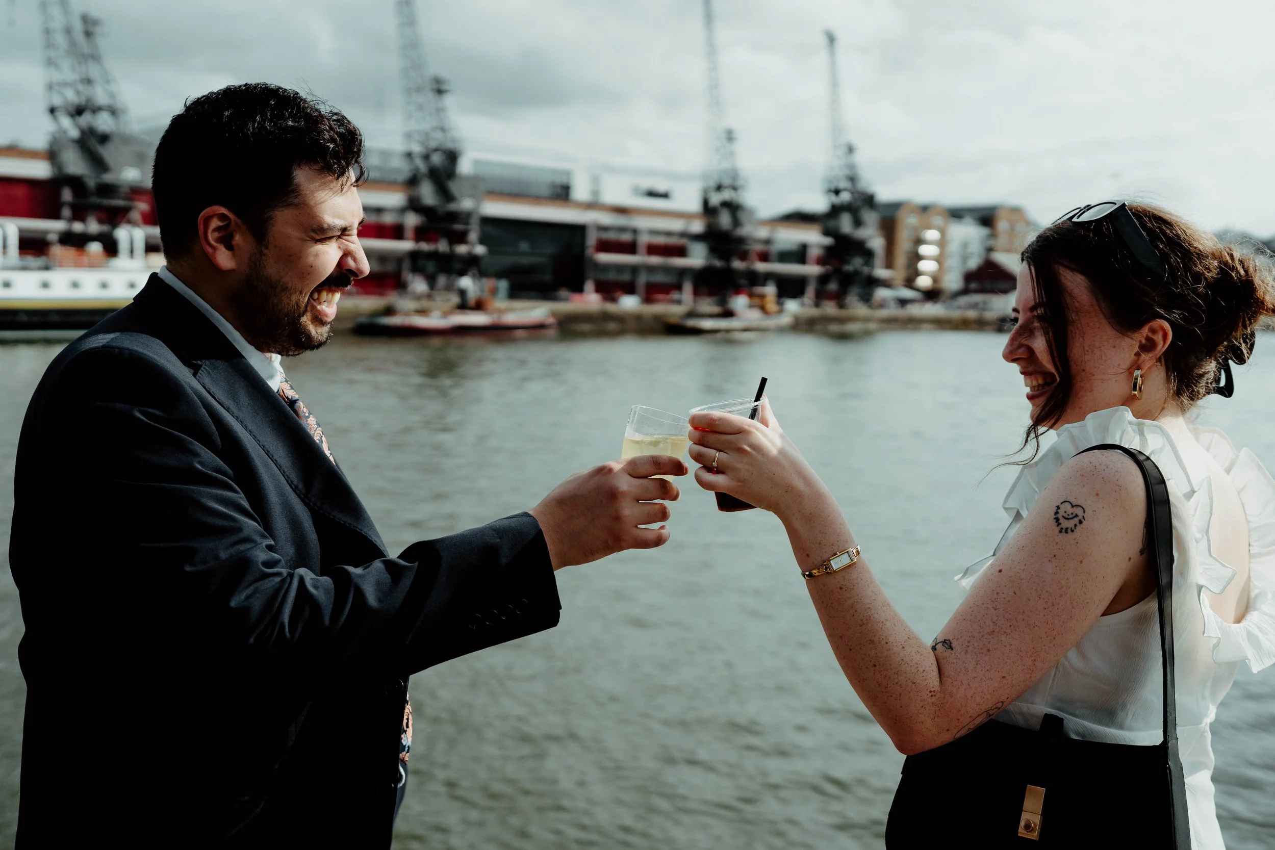 A man in a suit and a woman with tattoos and sunglasses on her head are smiling and laughing as they toast drinks by a waterfront with boats and buildings in the background.