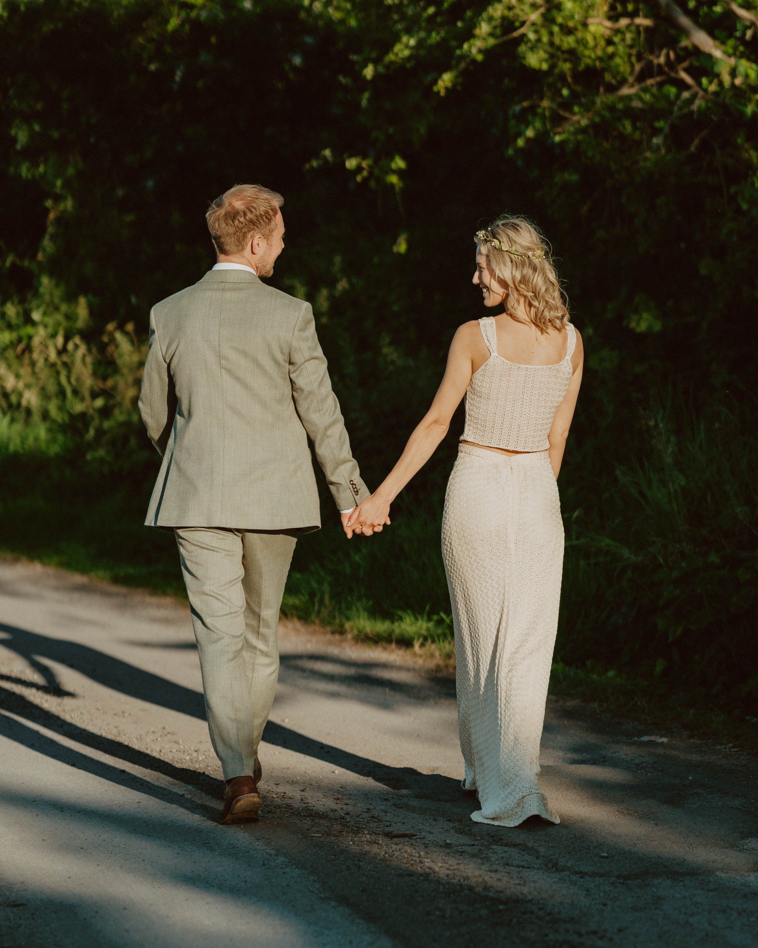 A couple holding hands and walking on a dirt path through a wooded area. The woman is wearing a light-colored two-piece dress and a floral headband, and the man is dressed in a light gray suit.