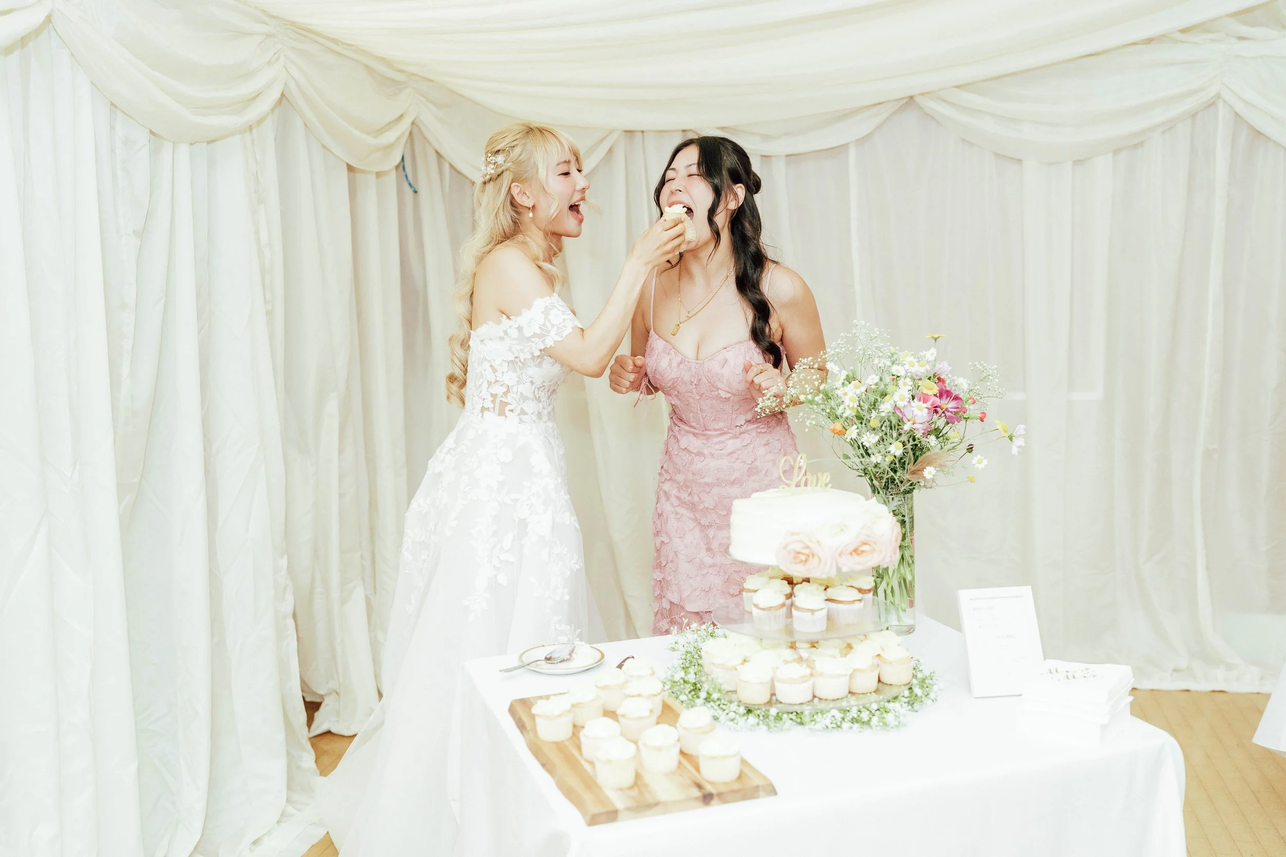 Two women at a wedding celebration, one in a white bridal gown and the other in a pink dress, sharing a moment while one feeds the other a piece of cake. There is a table with a layered cake, cupcakes, flowers, and a cake topper reading 'Love' in fro