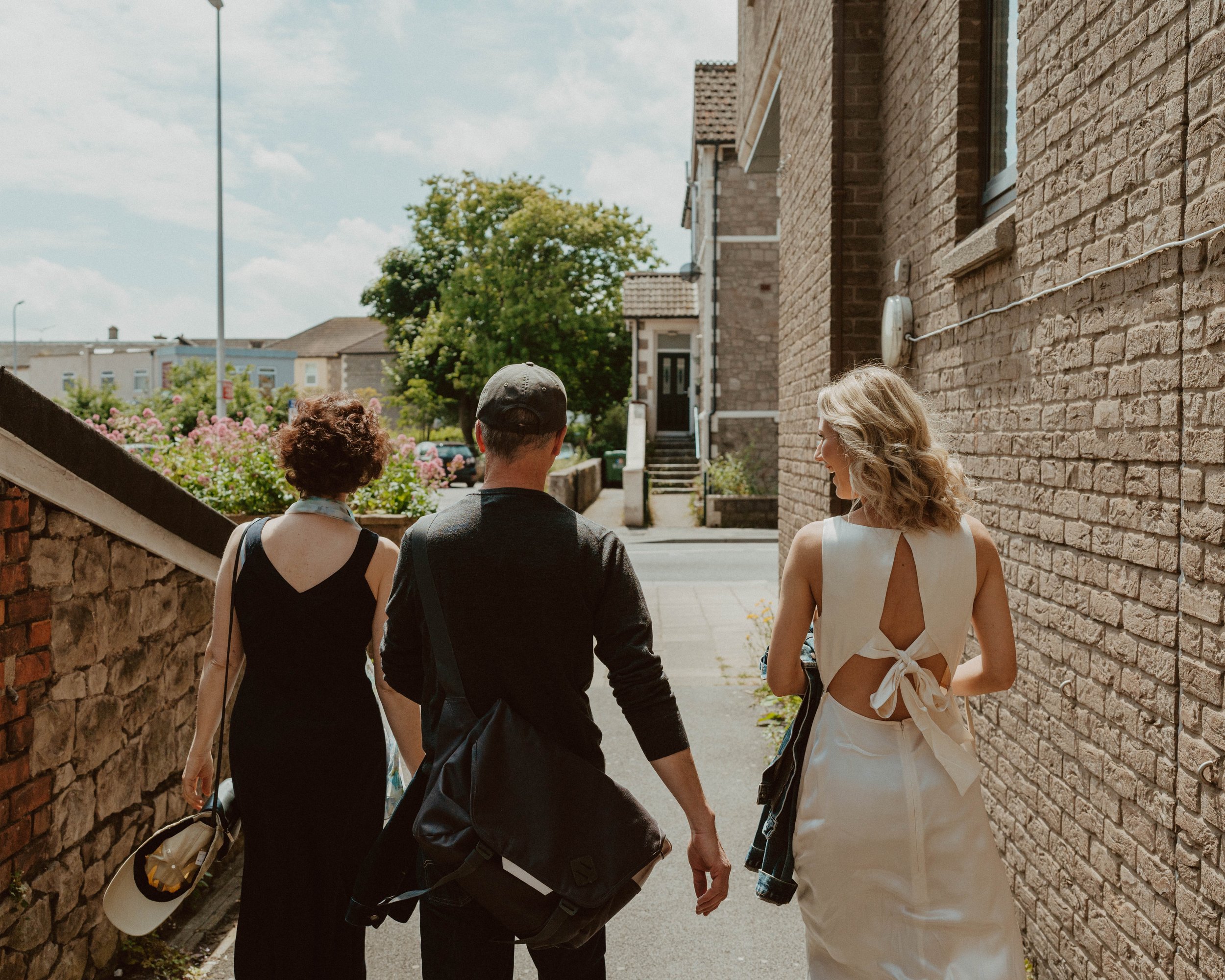 Three people walk down a city sidewalk on a sunny day, two women and a man, with houses and greenery in the background.
