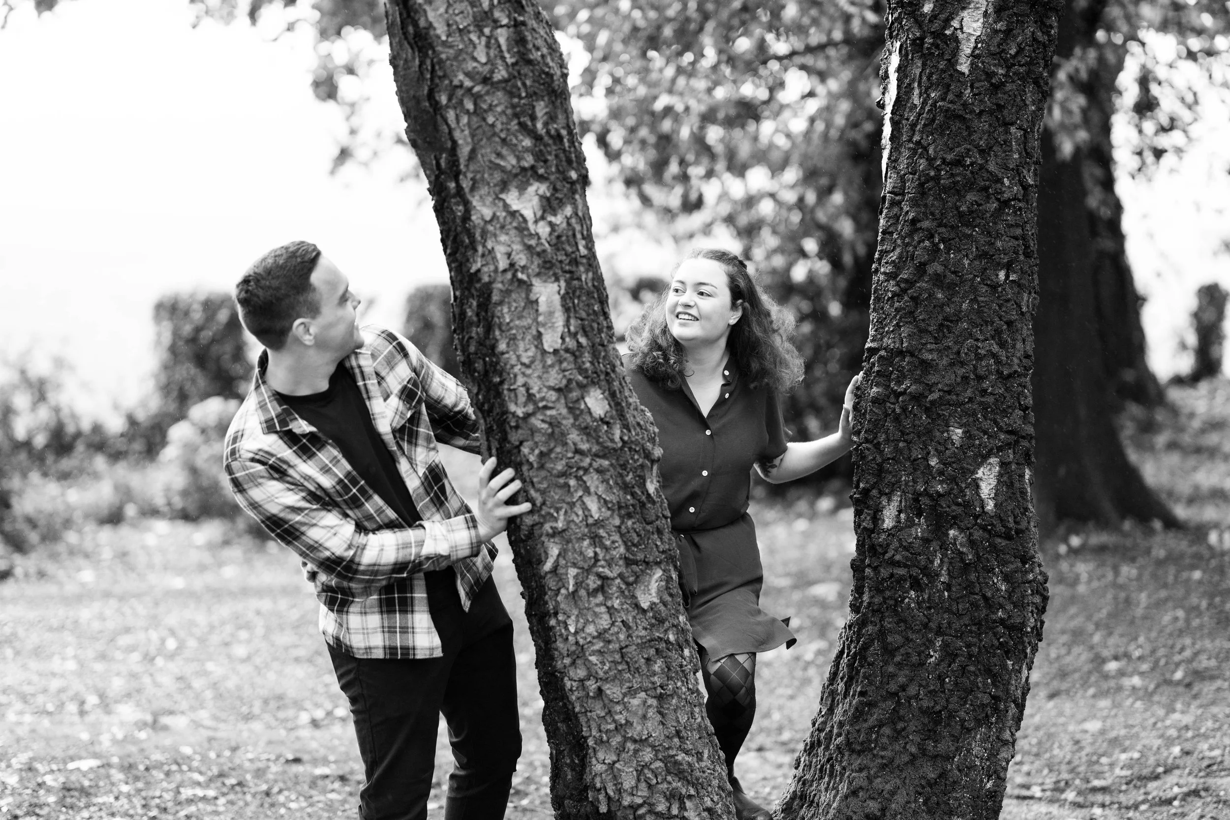 A black and white photo of a man and woman playing hide and seek behind trees in a park.