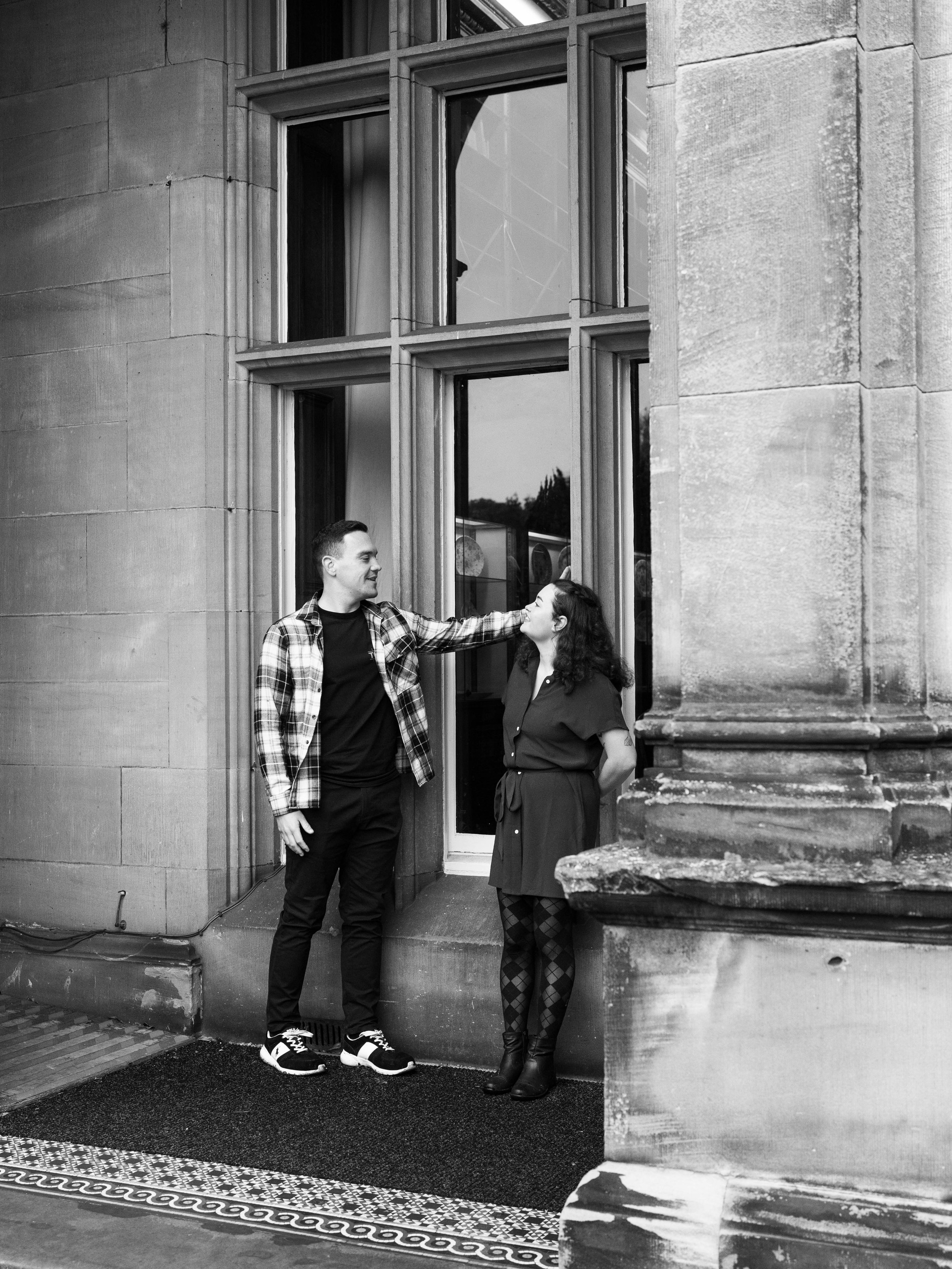 A black-and-white photo showing a man and woman standing outside a building, engaging in conversation. The man is touching the woman's shoulder, and they are facing each other. The building has large windows and stone walls.