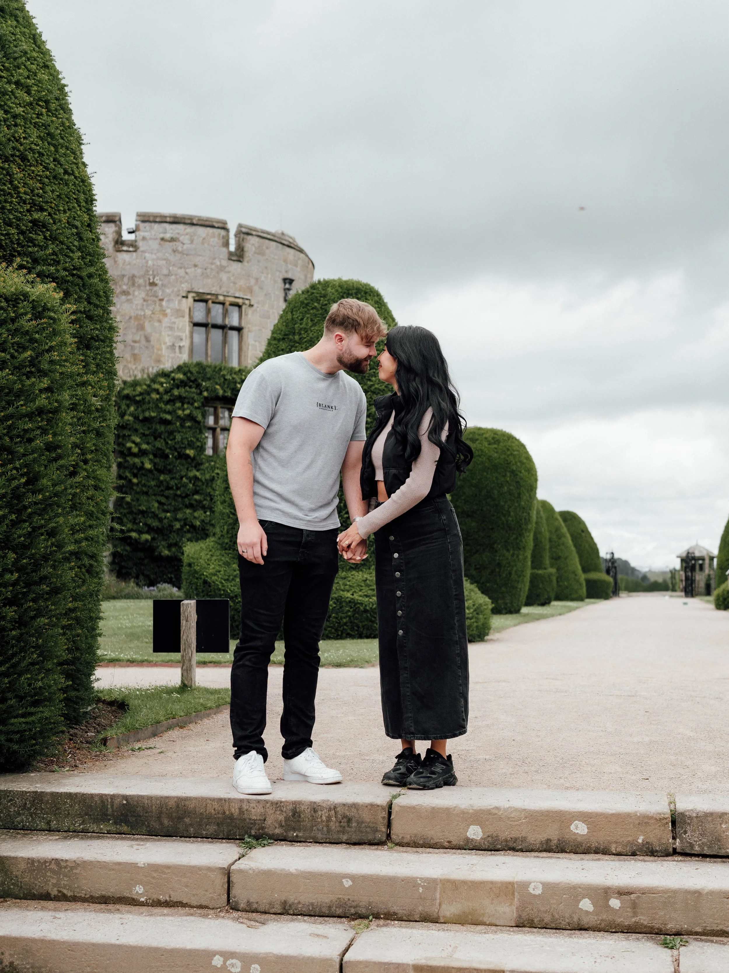 A couple standing on stone steps in a garden, nose to nose, holding hands, with a castle-like building and trimmed bushes in the background.