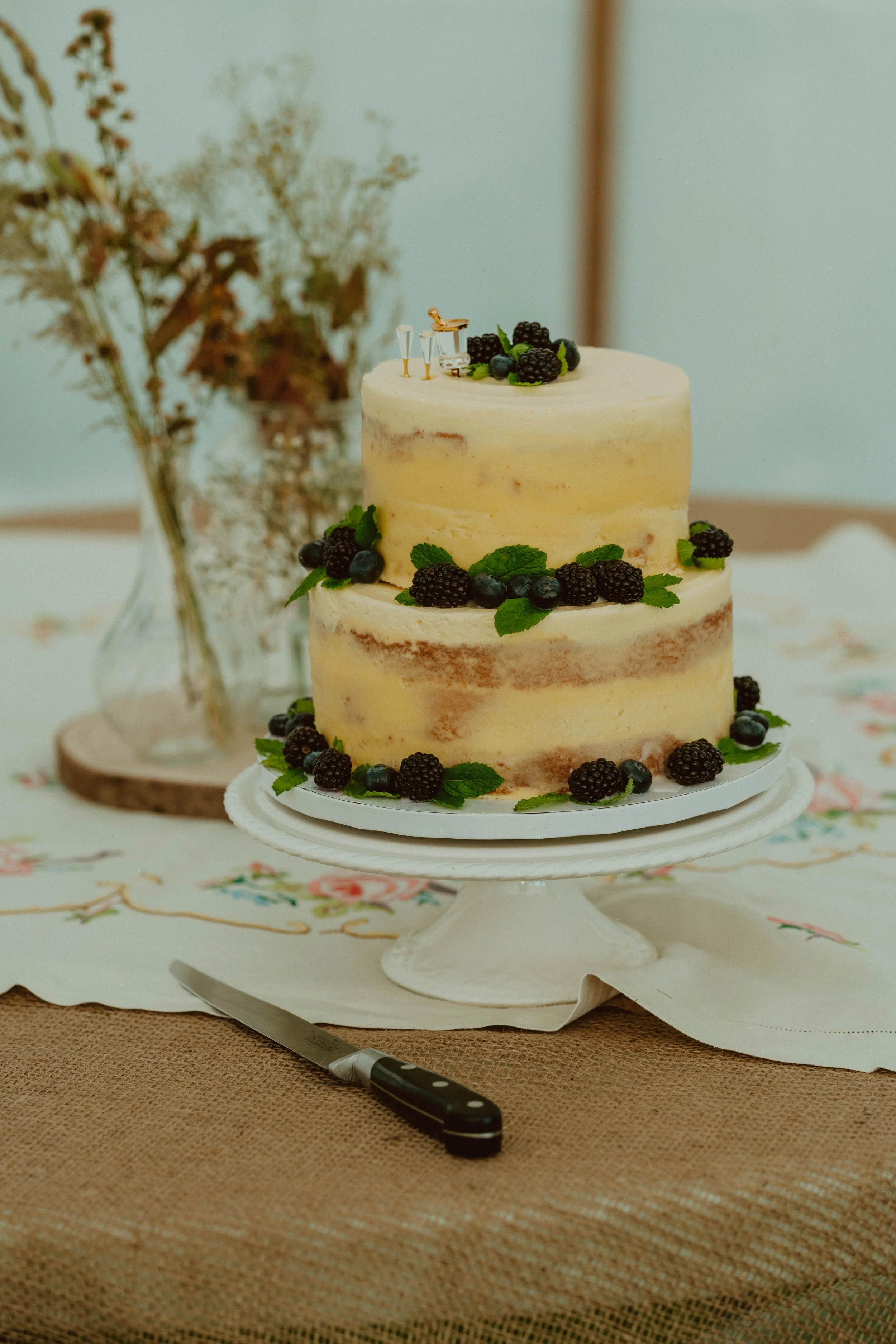A two-tiered naked wedding cake decorated with blackberries, blueberries, and mint leaves, placed on a white cake stand on a table with floral tablecloth and a serving knife nearby.