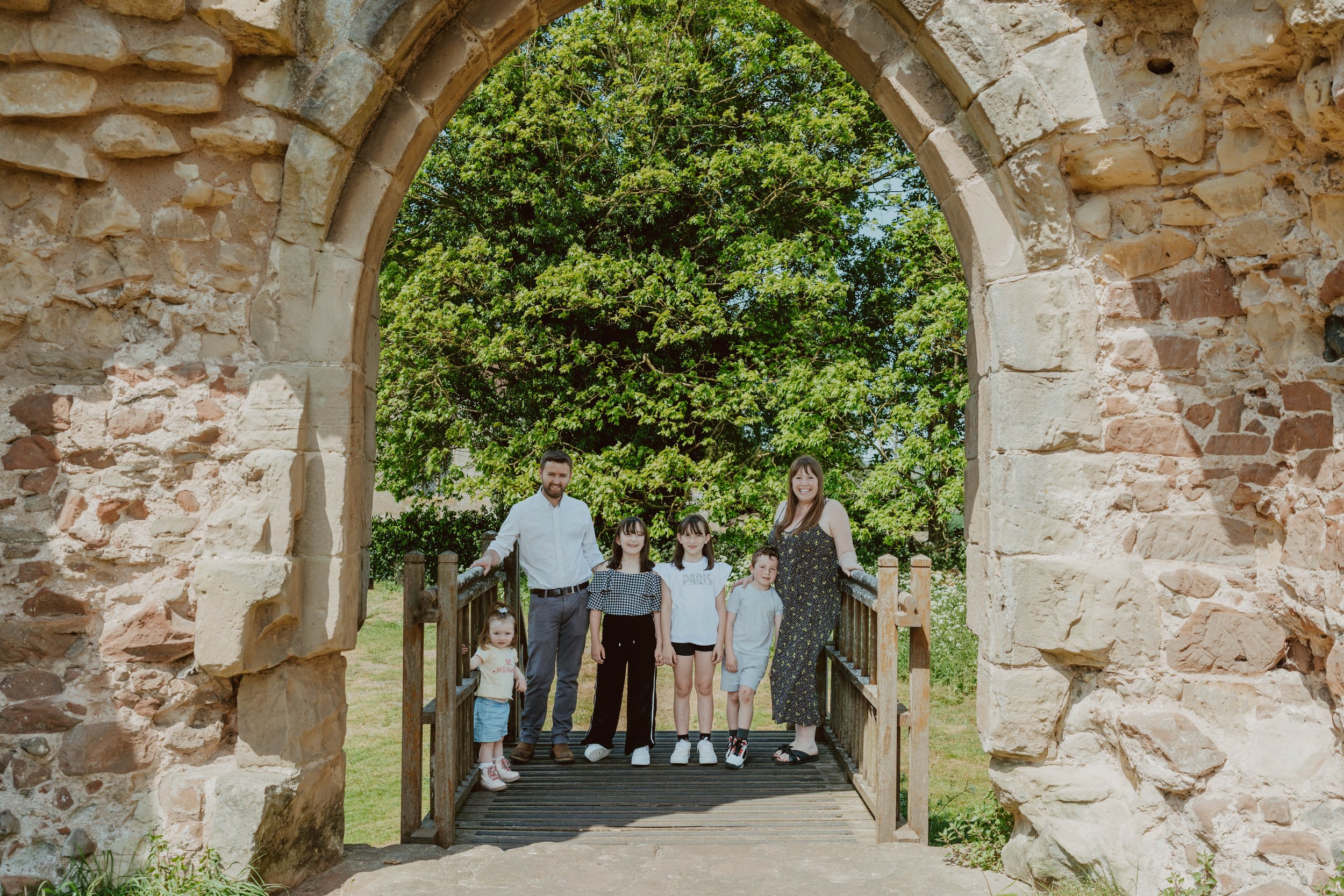 Family of five standing on a small wooden bridge under a stone archway, with lush green trees in the background.