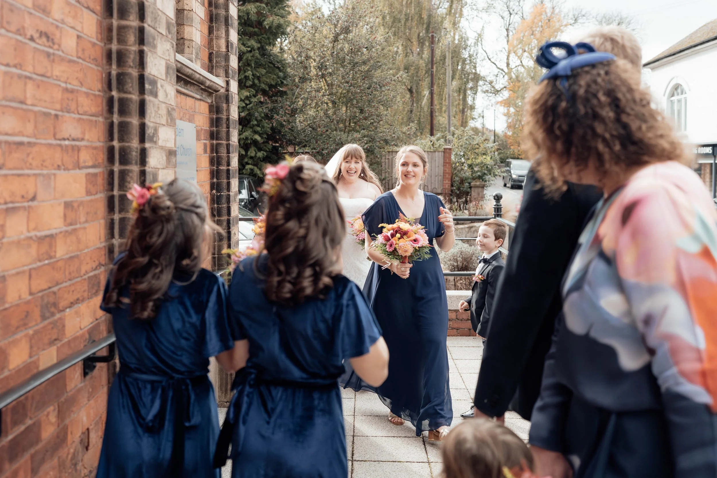 A bride in a white wedding dress is walking outside with bridesmaids and flower girls, all holding bouquets, near a brick building with trees in the background.