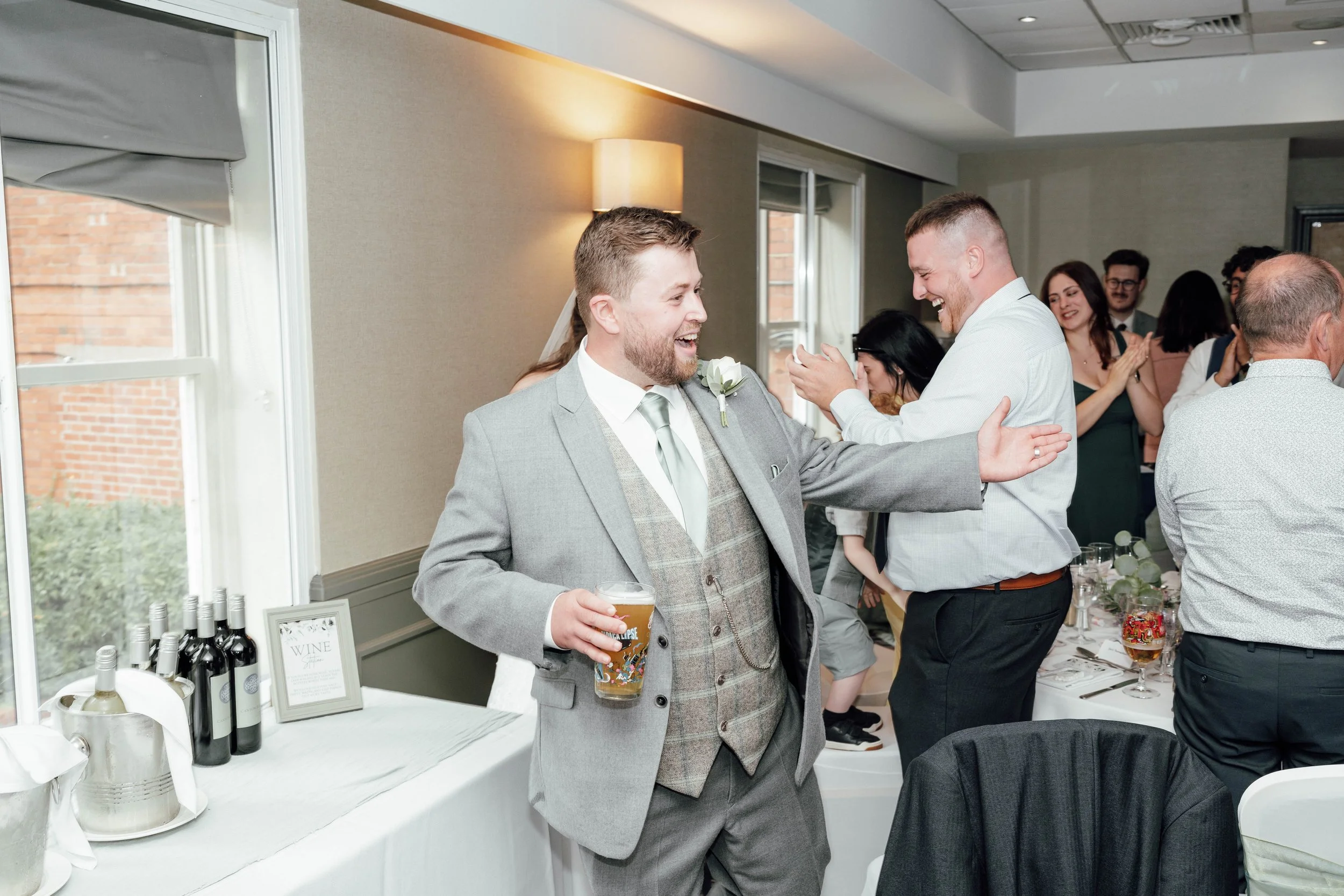 Two men celebrating at a wedding reception, one in a gray suit with a drink and the other in a white shirt laughing, with guests clapping in the background.