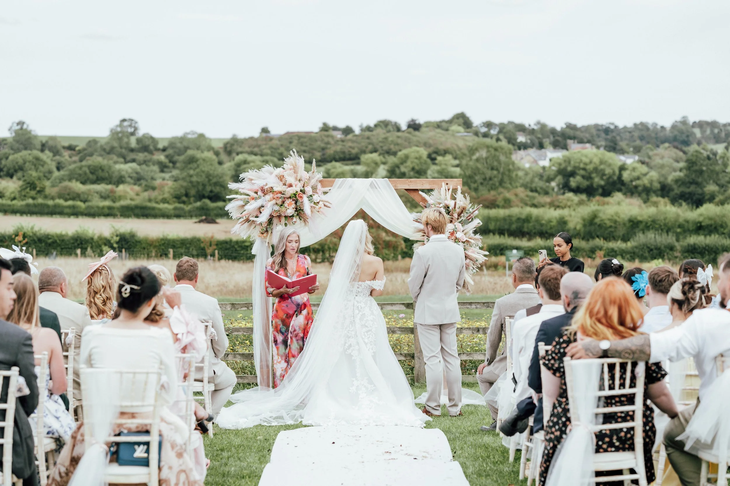 A wedding ceremony taking place outdoors on a grassy field with a lush green landscape in the background. The bride and groom stand under a wooden arch decorated with white drapery and large floral arrangements. The officiant is reading from a book, 