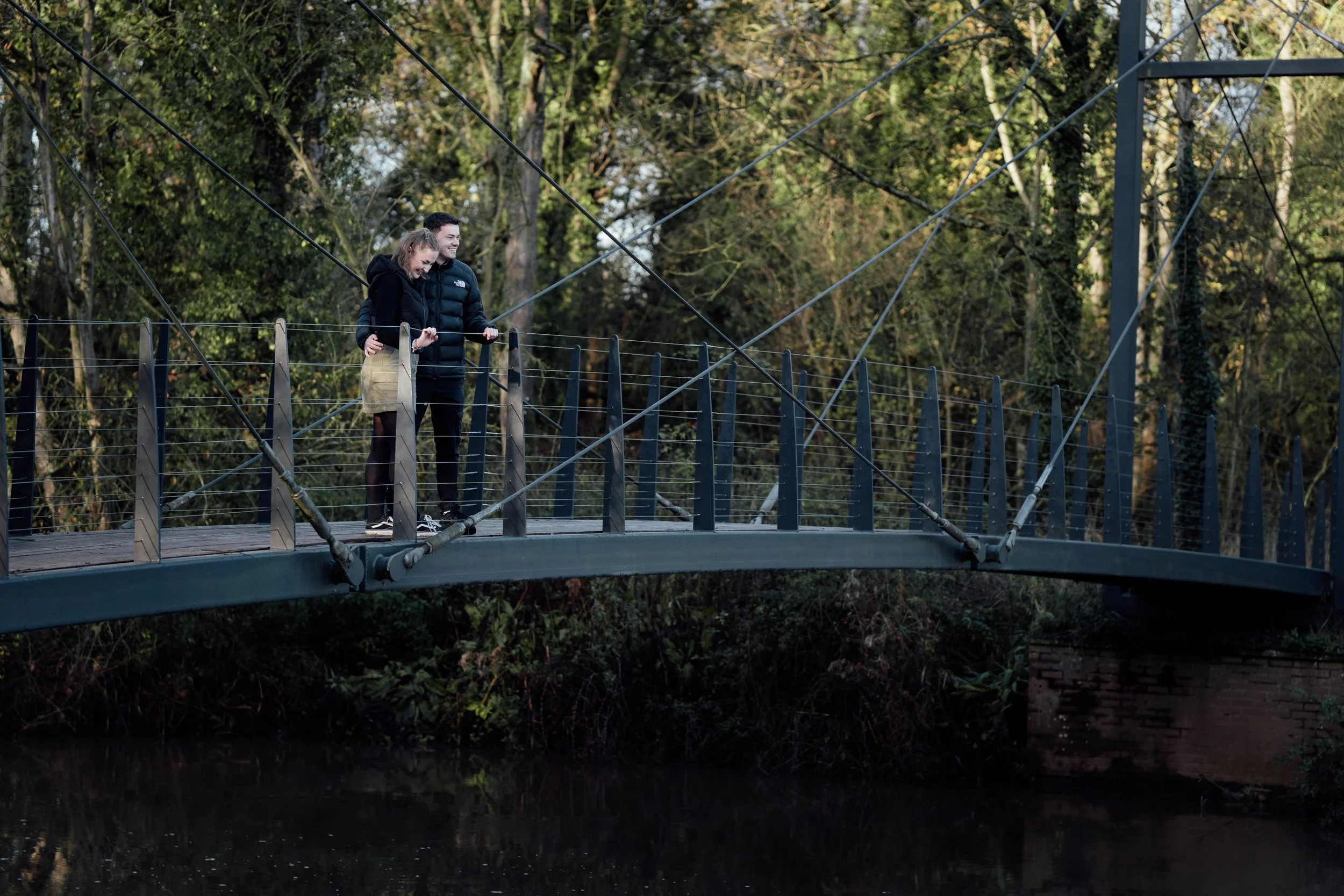 A couple stands on a suspension bridge in a wooded area, smiling and enjoying the view.