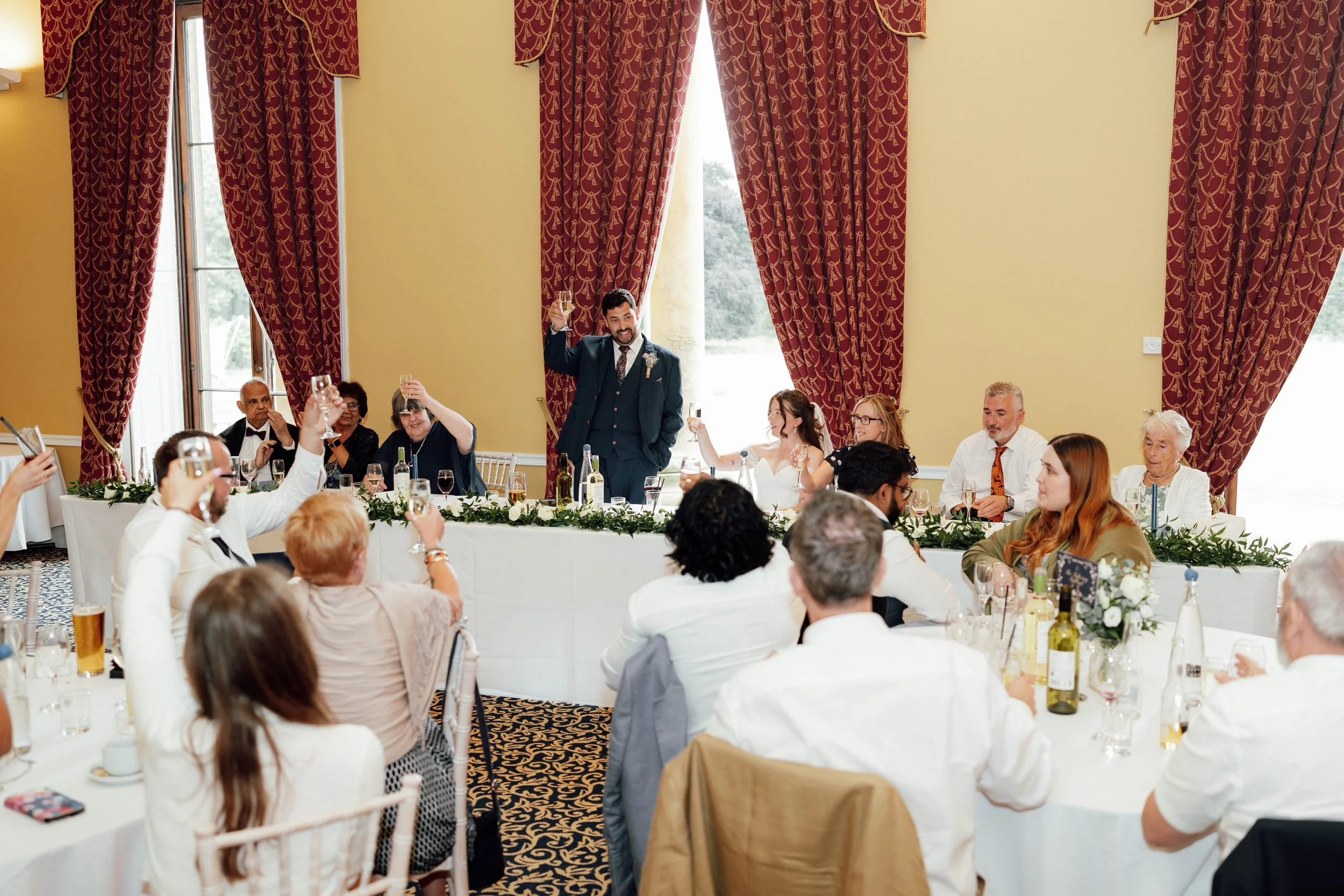 A group of people at a wedding reception raising glasses in a toast, with the bride and groom at the head table in an elegant room with large windows and red curtains.