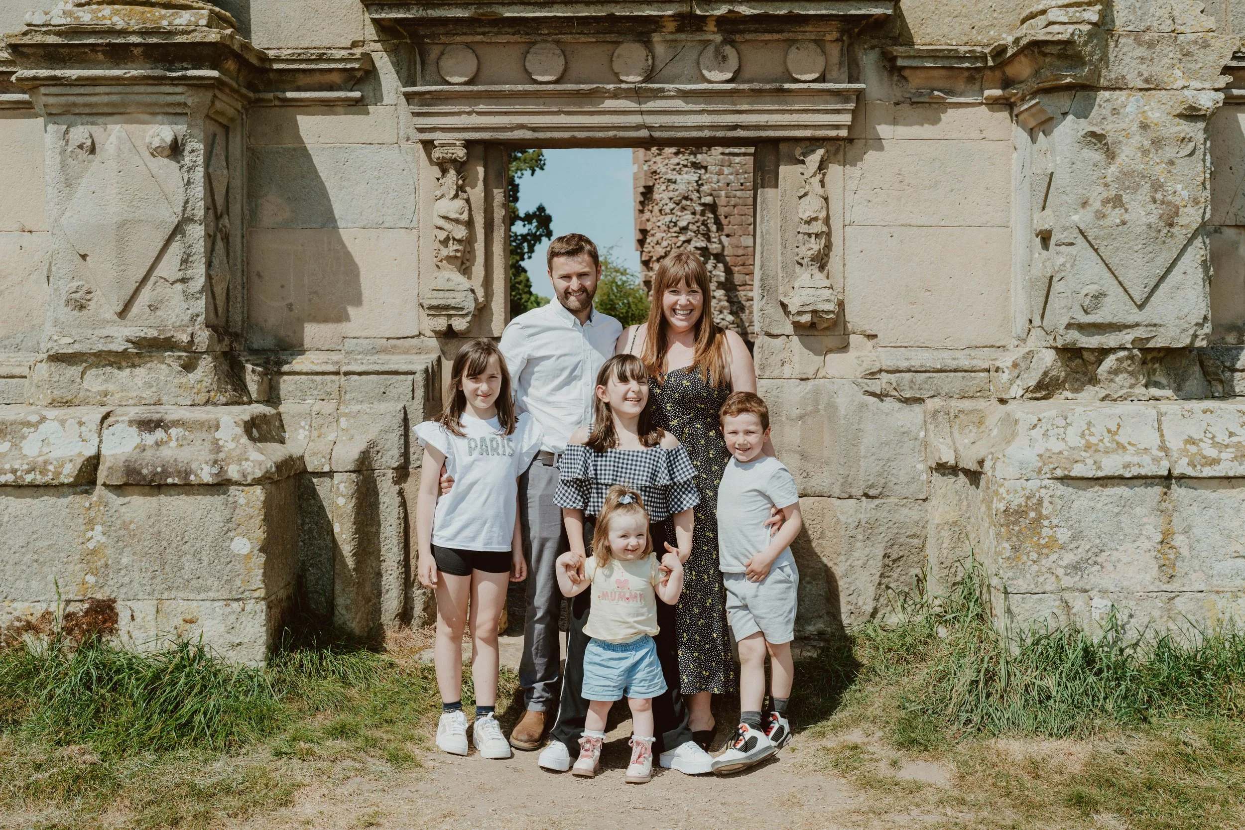 Family of seven standing under a stone archway with ancient ruins in the background on a sunny day.