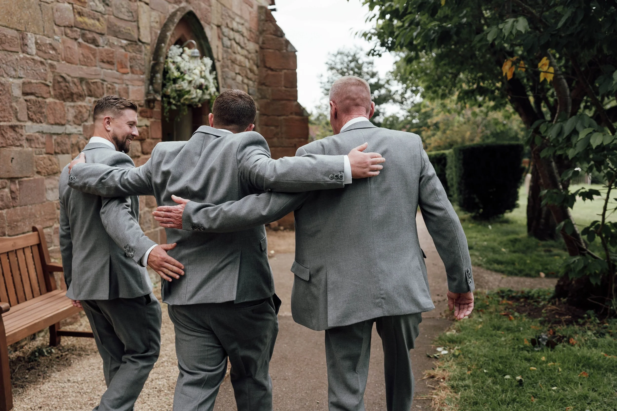 Four men in gray suits walking together outdoors, with one arm around each other's shoulders, near a brick building and surrounded by greenery.