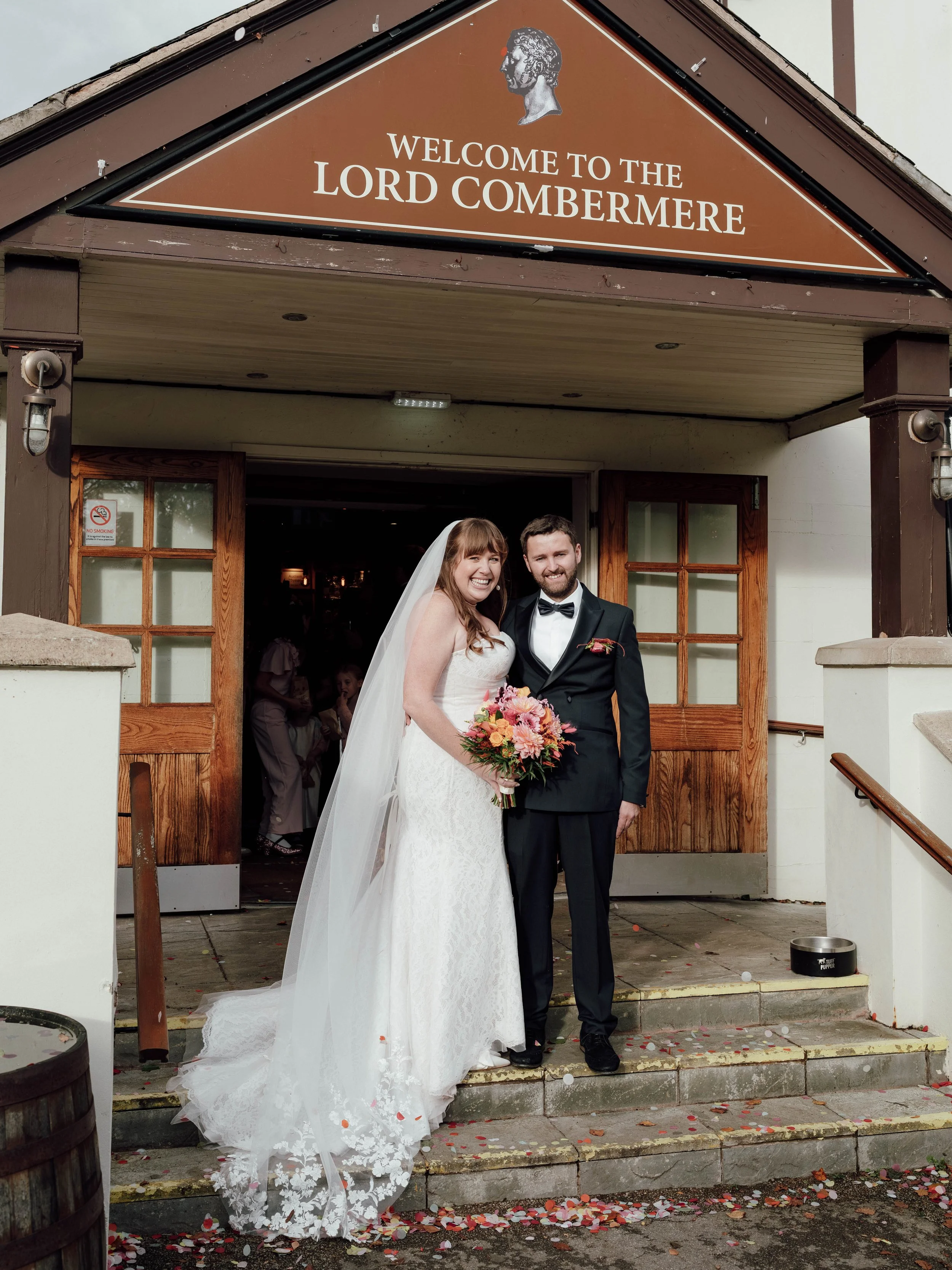 Bride and groom standing on church steps after wedding, smiling, bride holding bouquet of pink flowers, wedding attire, confetti on ground.